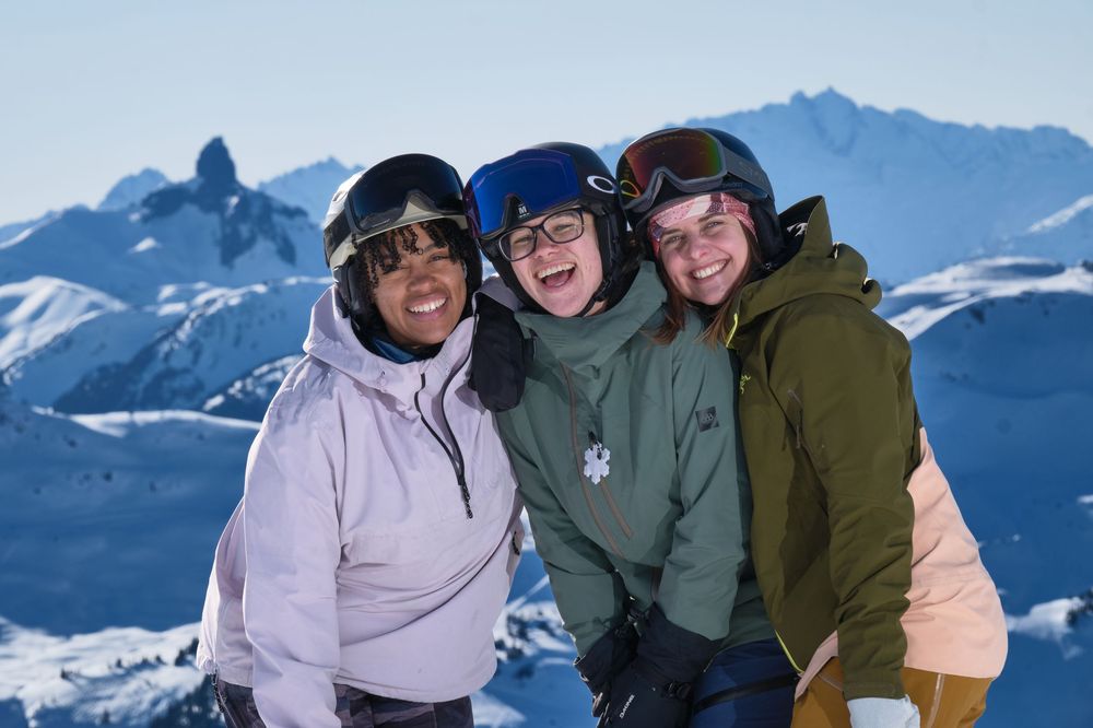 Three friends in ski gear smile together on a snowy mountain with snow-capped peaks in the background.