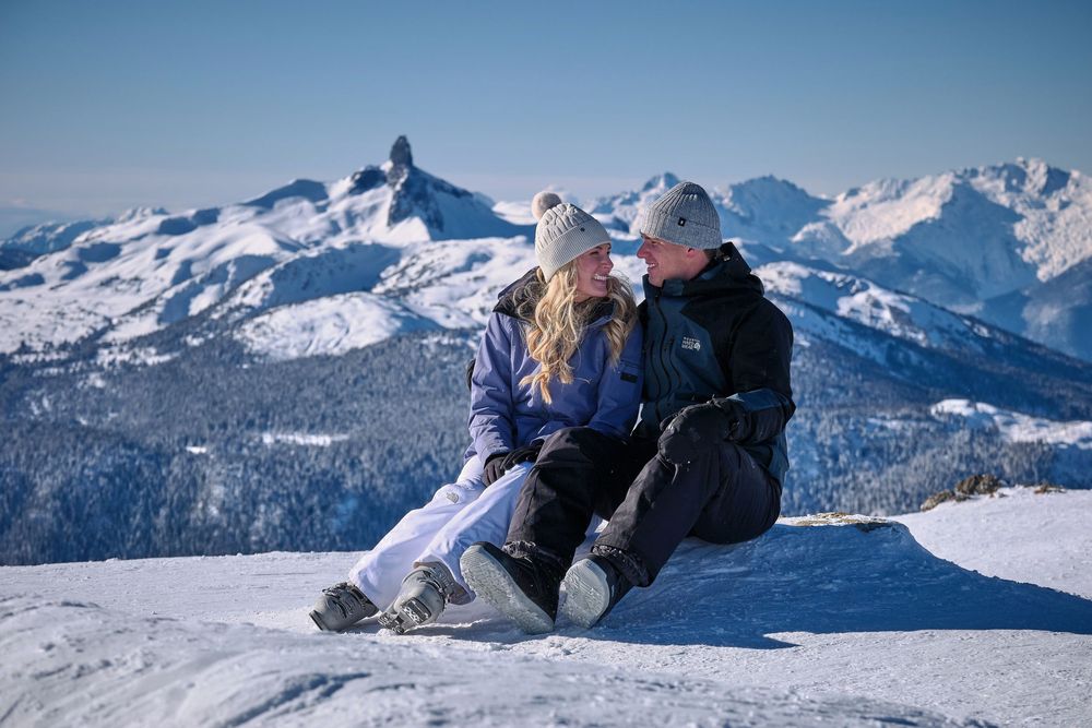 Two skiers sitting together on snowy mountain peak with scenic winter mountain landscape and blue sky in background.