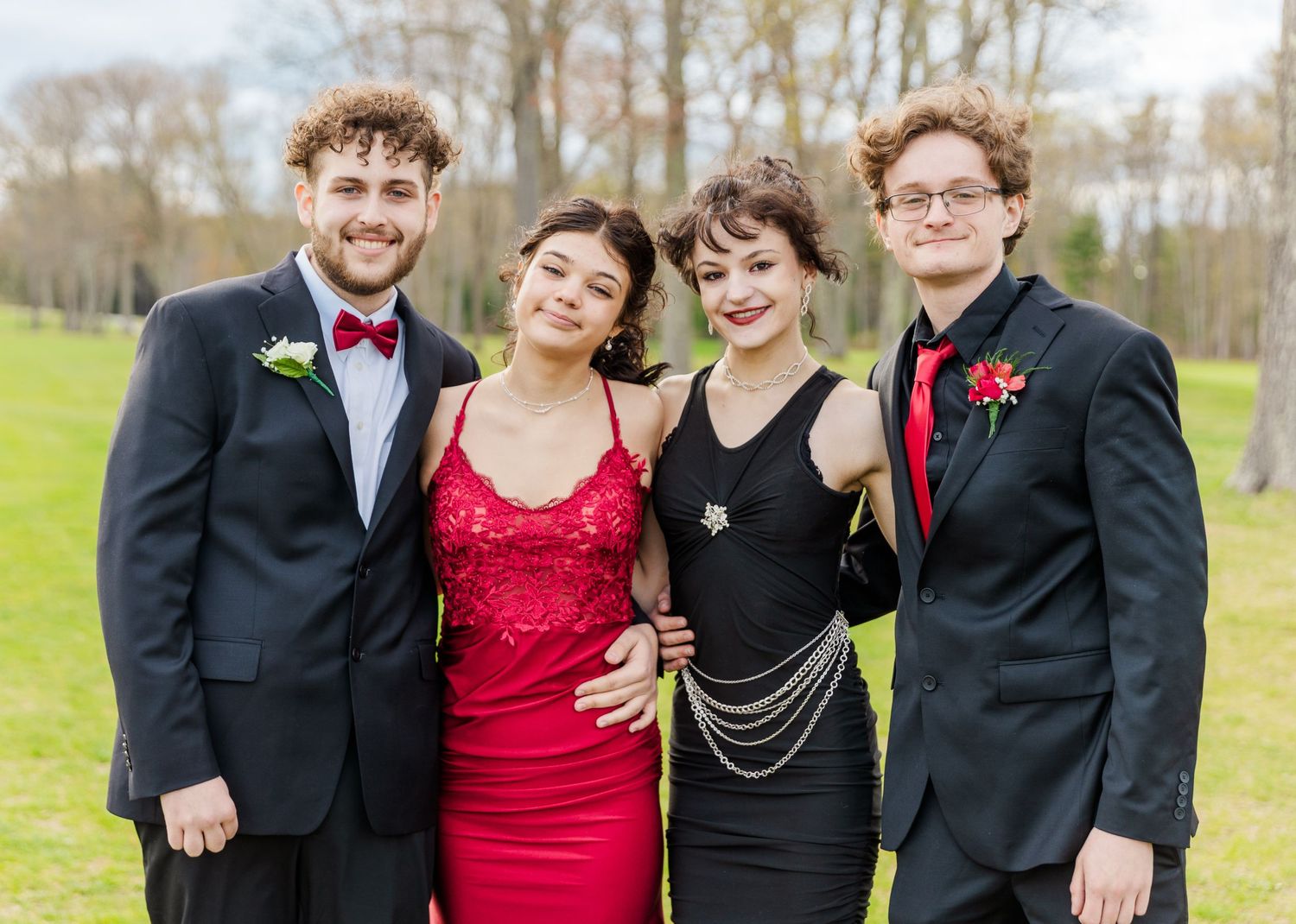 A group in formal attire poses together outdoors, with two in suits and red ties and two in elegant dresses.