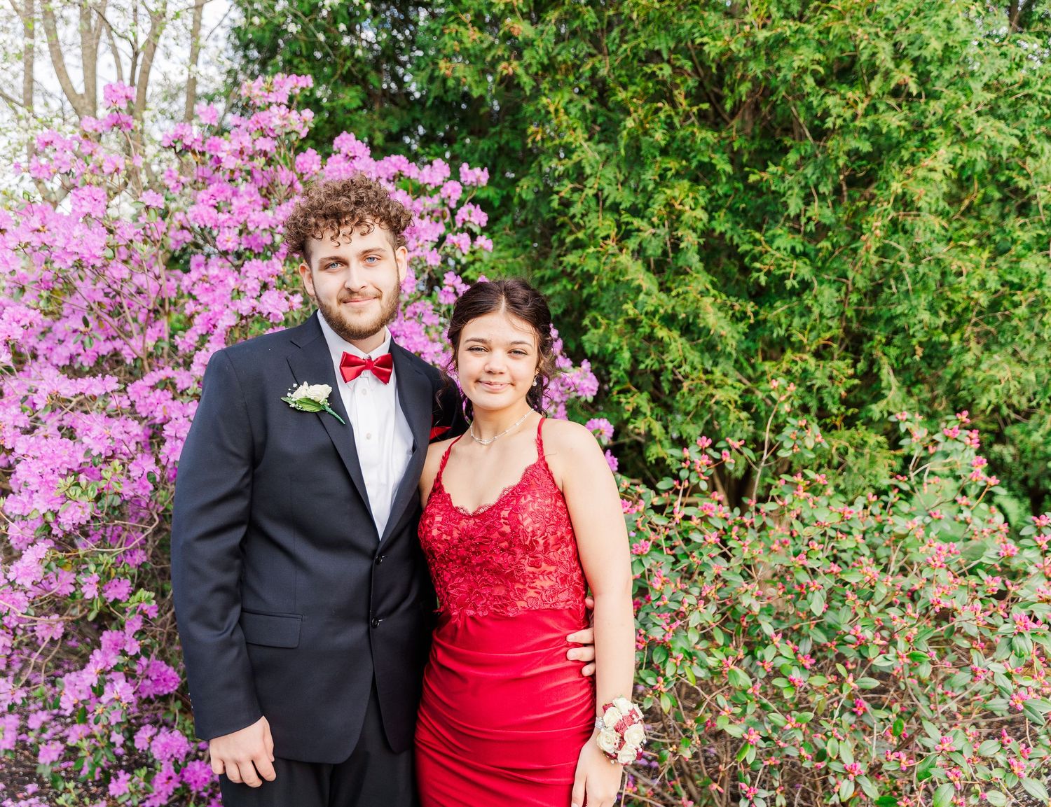 A couple poses in formal wear in front of blooming pink azalea bushes.