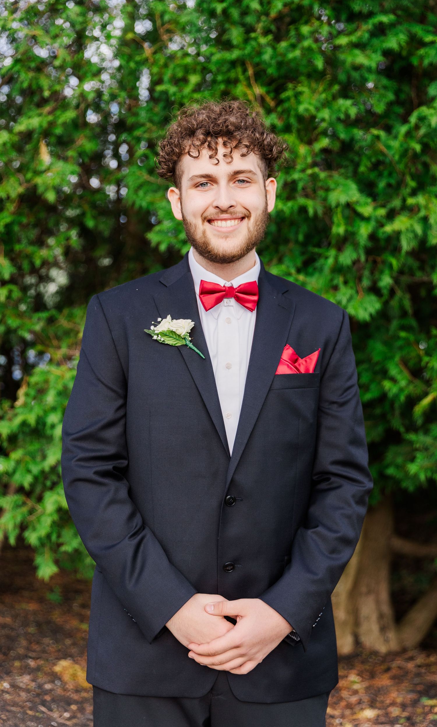 A person in a black suit with red bowtie and boutonniere smiles at the camera.