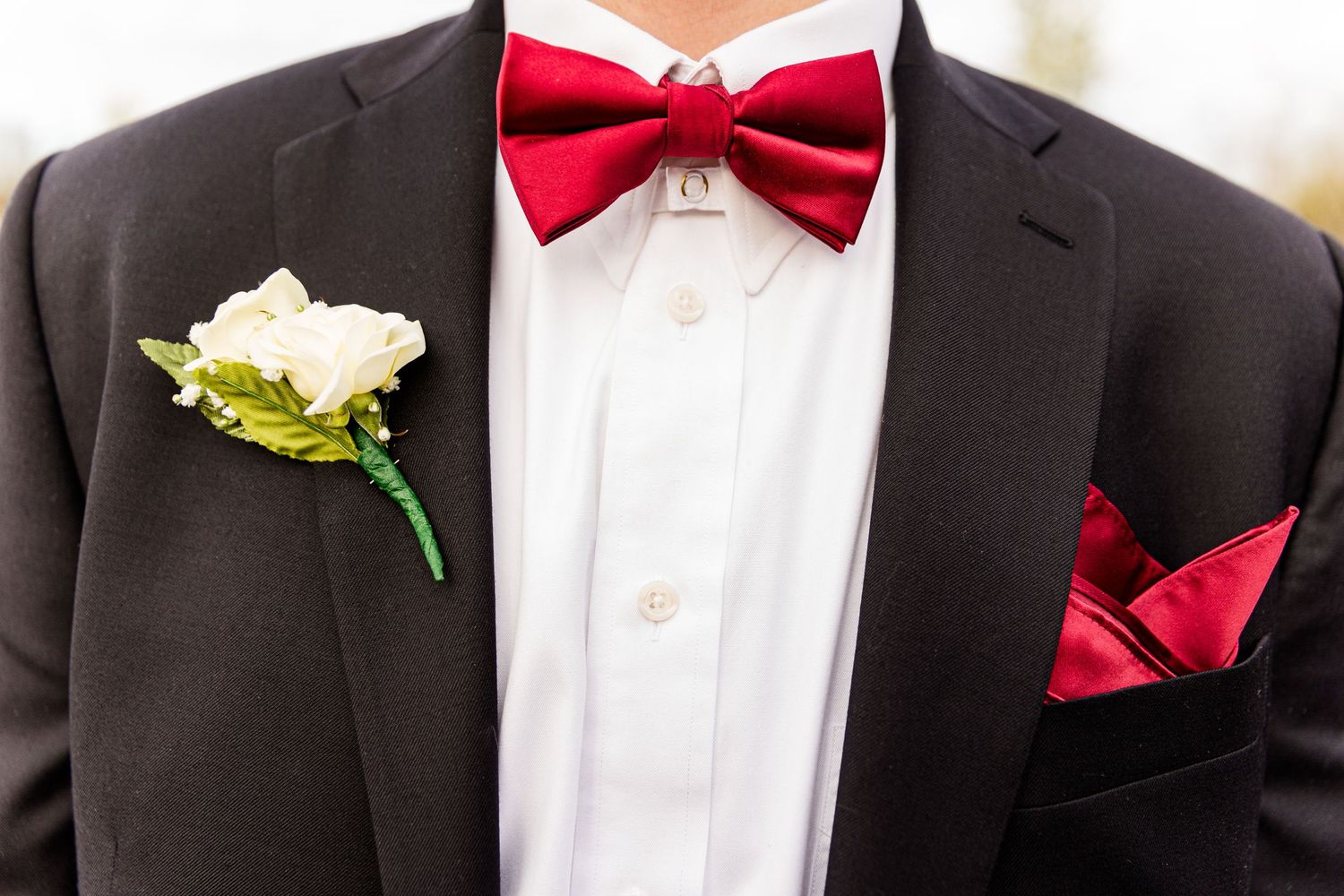 A close-up of a black suit with red bowtie, white boutonniere, and red pocket square.