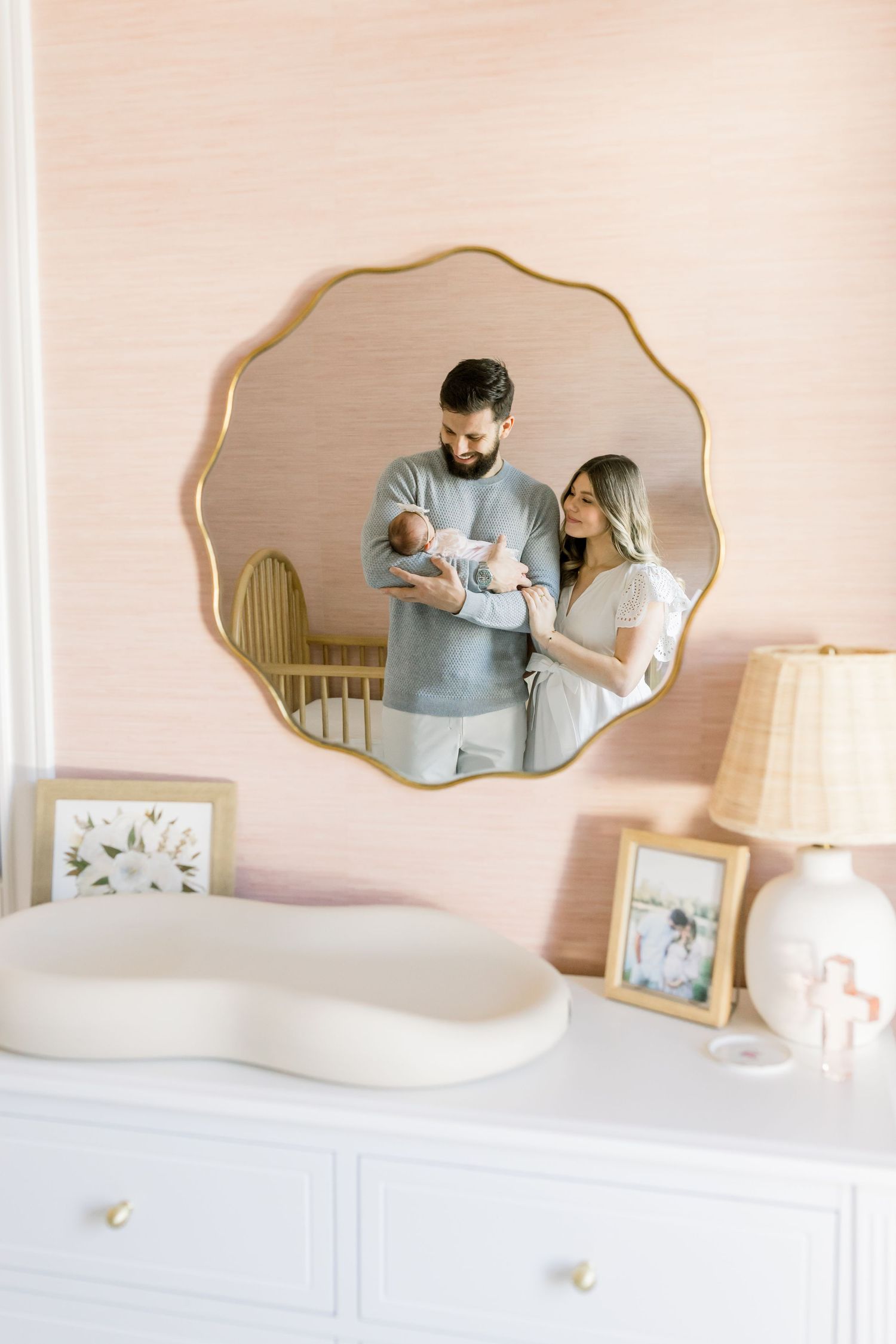 Mirror reflection captures tender family moment with newborn in pink-walled nursery with decorative details.