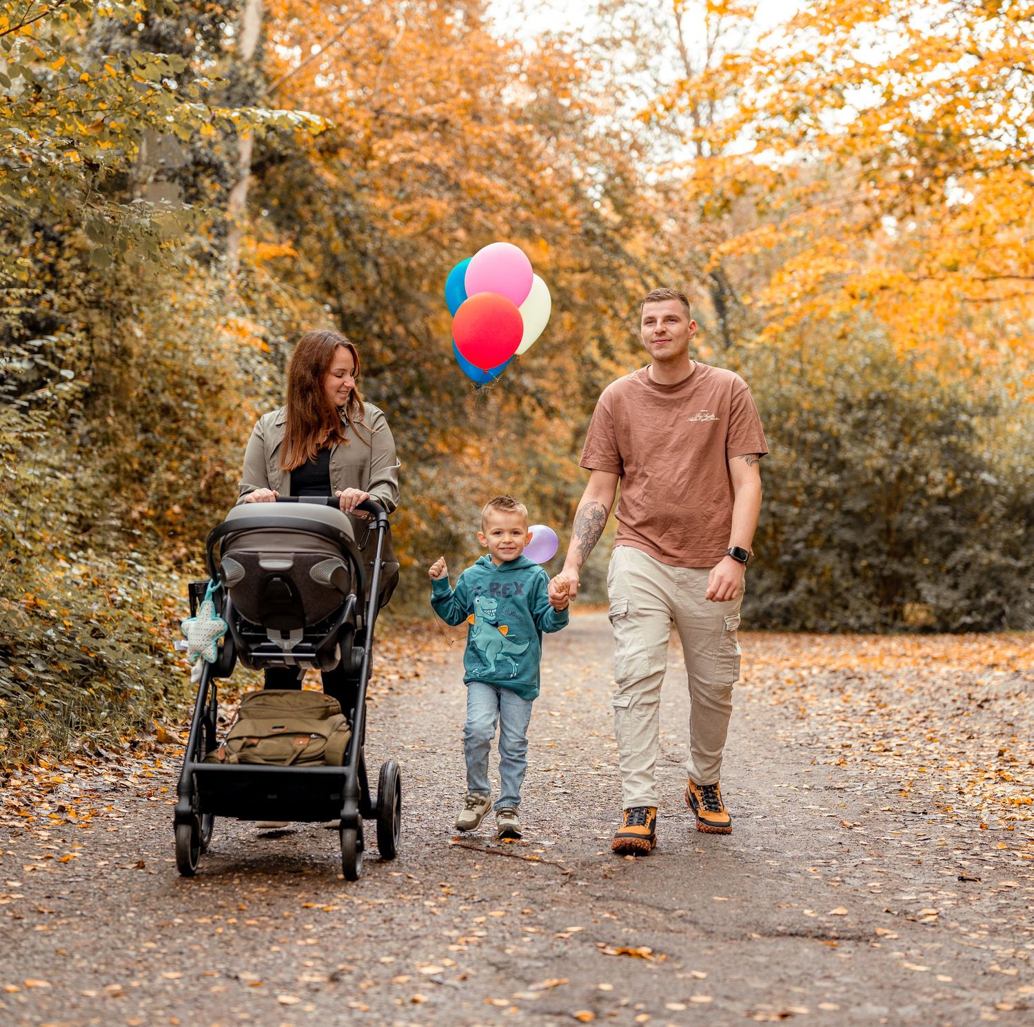 Family walking on autumn trail with stroller and red balloon surrounded by golden fall foliage.
