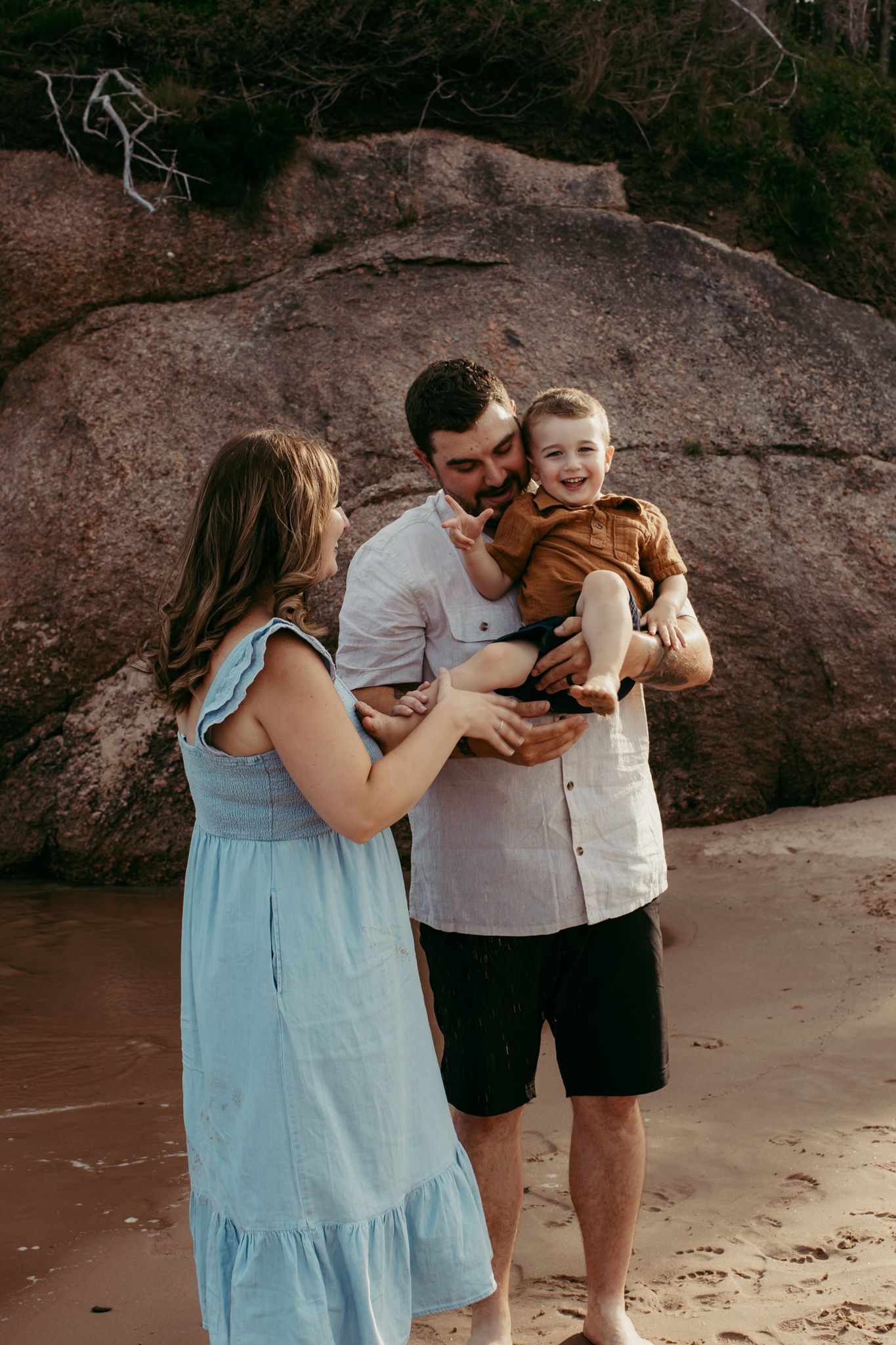 Family sharing playful moment together on beach with large rock formation in background.