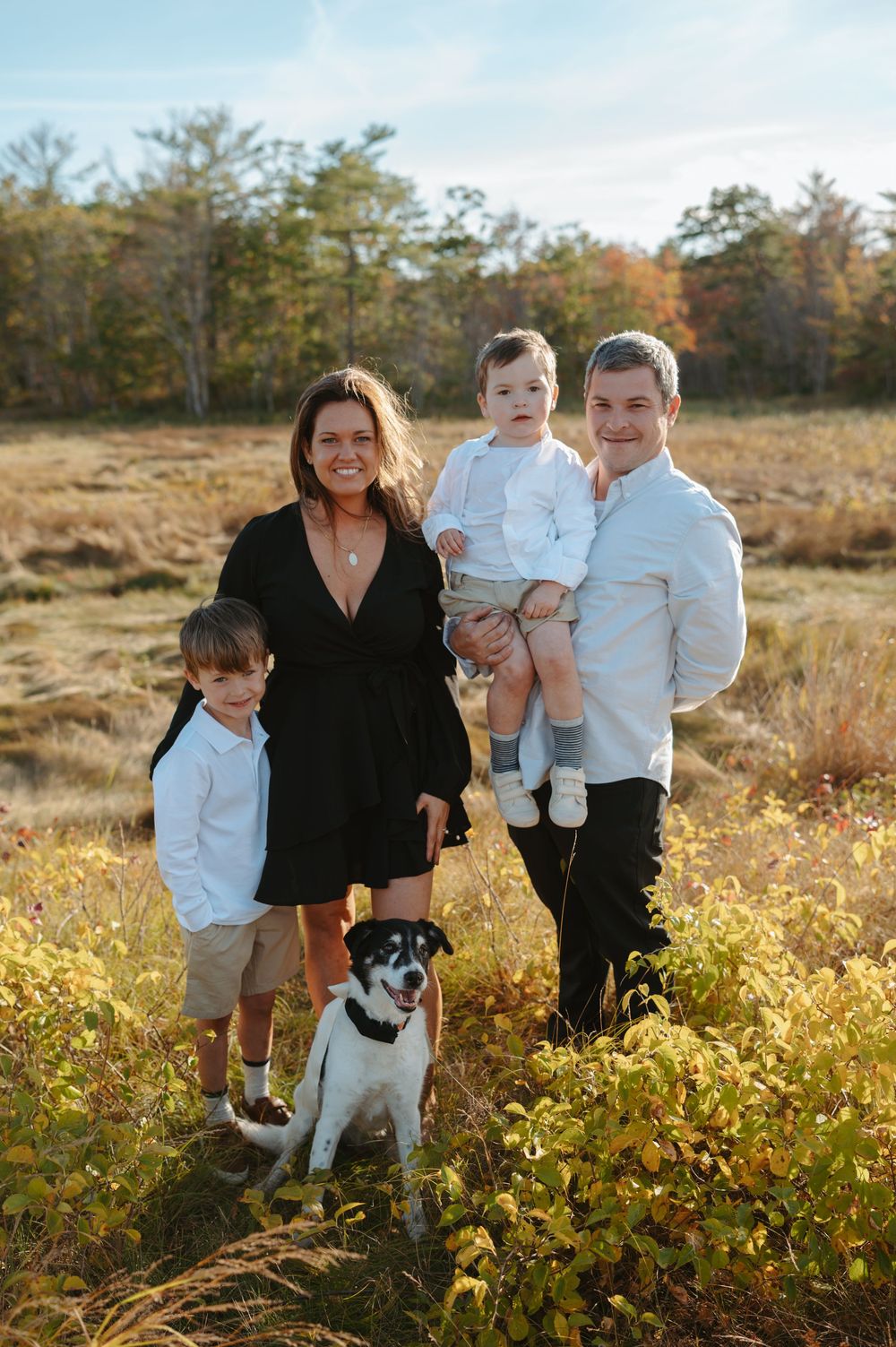 Family and their black and white dog pose together in a sunny autumn field with golden vegetation.