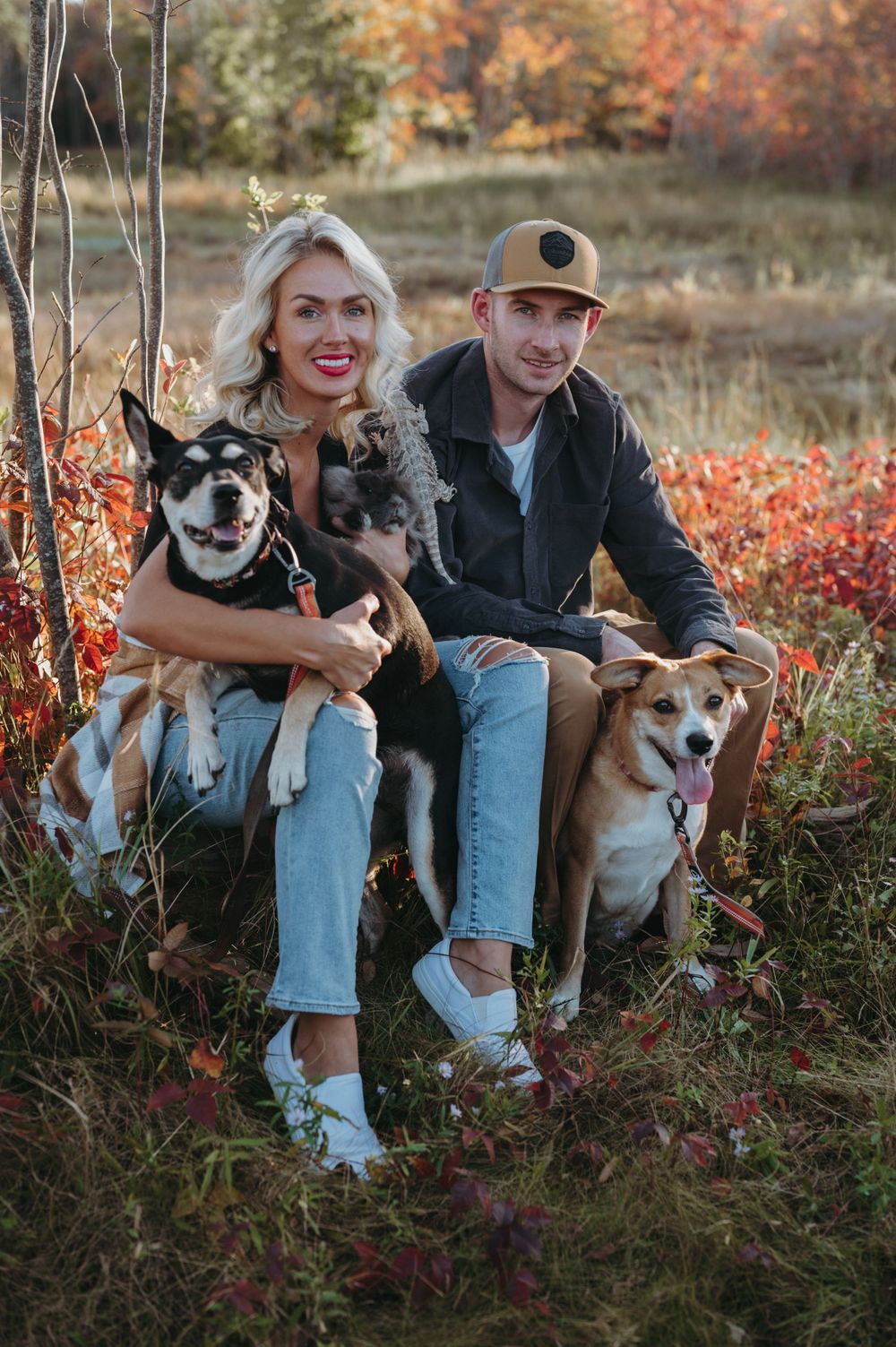 Happy couple sitting outdoors with their two smiling dogs surrounded by fall foliage.