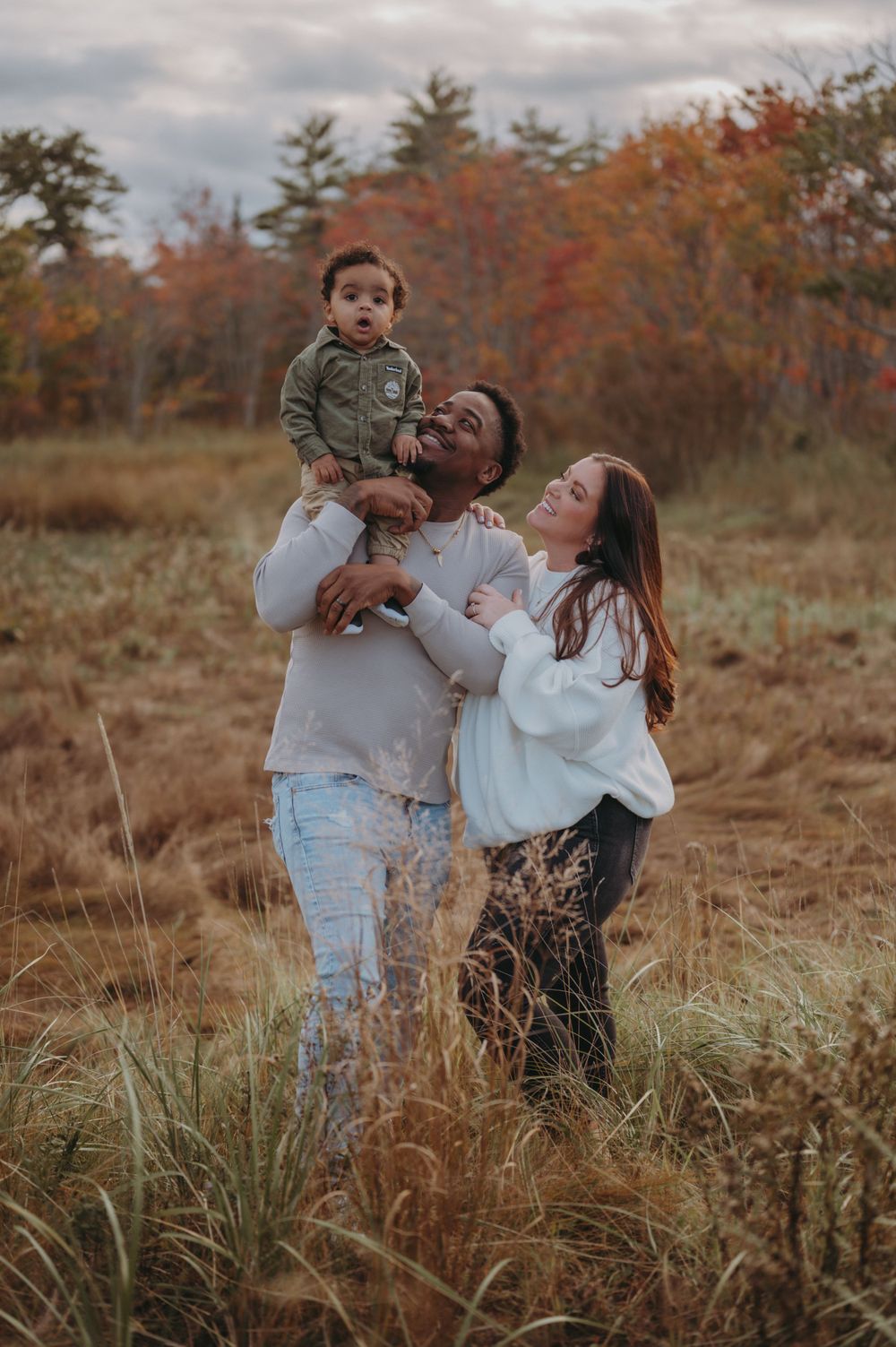 A joyful family plays together in a field surrounded by fall foliage.