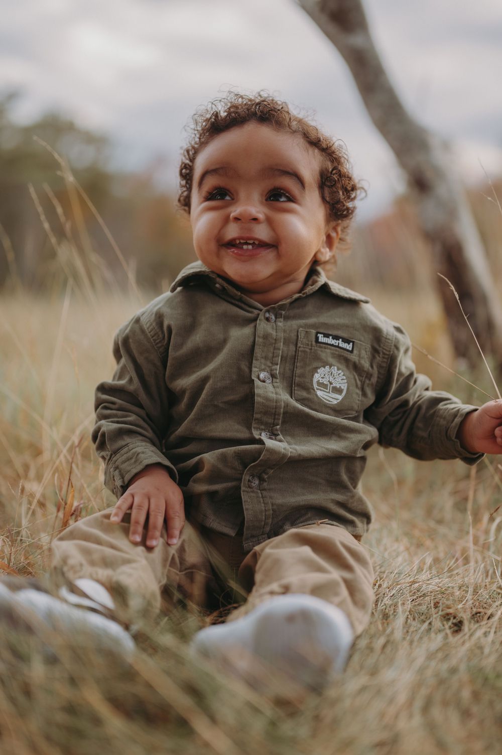 A toddler in a brown hoodie sits in tall grass with a bright smile.
