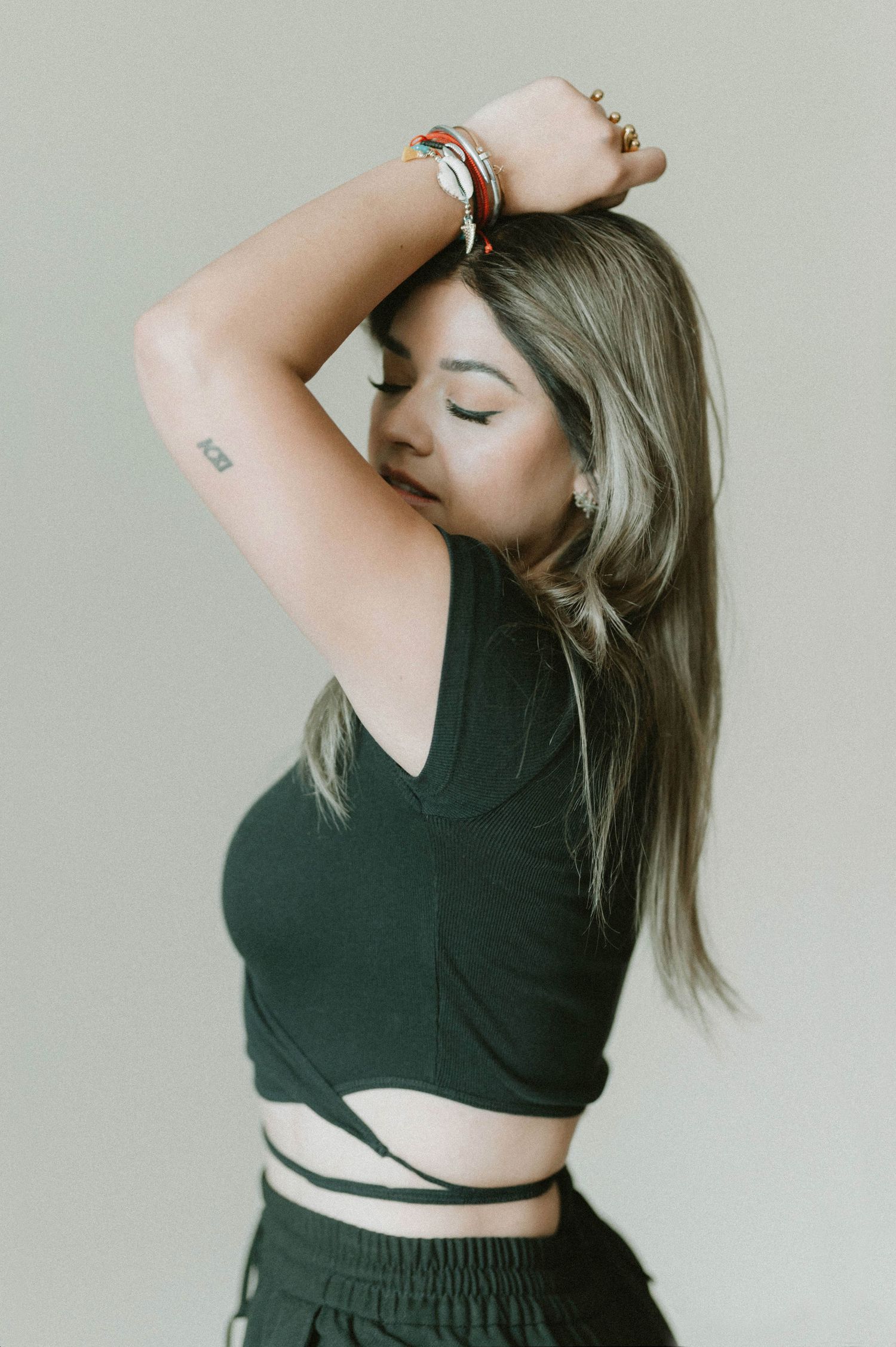 A fashion model in a black crop top poses against a neutral background with bracelets and flowing hair.