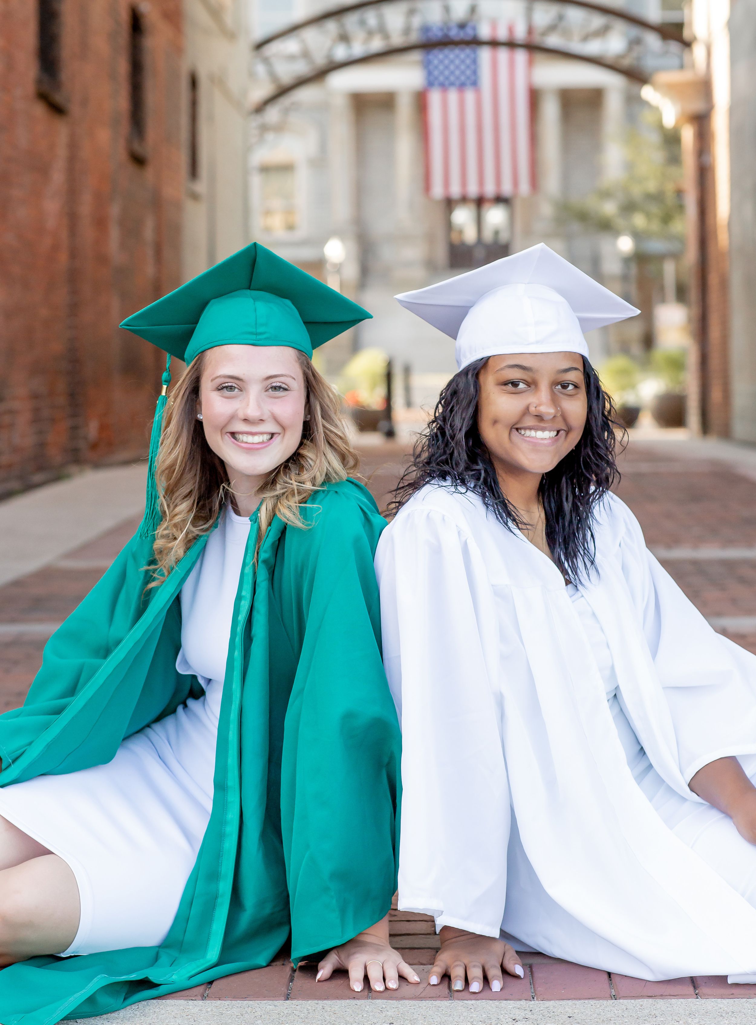 Two smiling graduates in green and white cap and gown sitting on brick path with American flag backdrop.