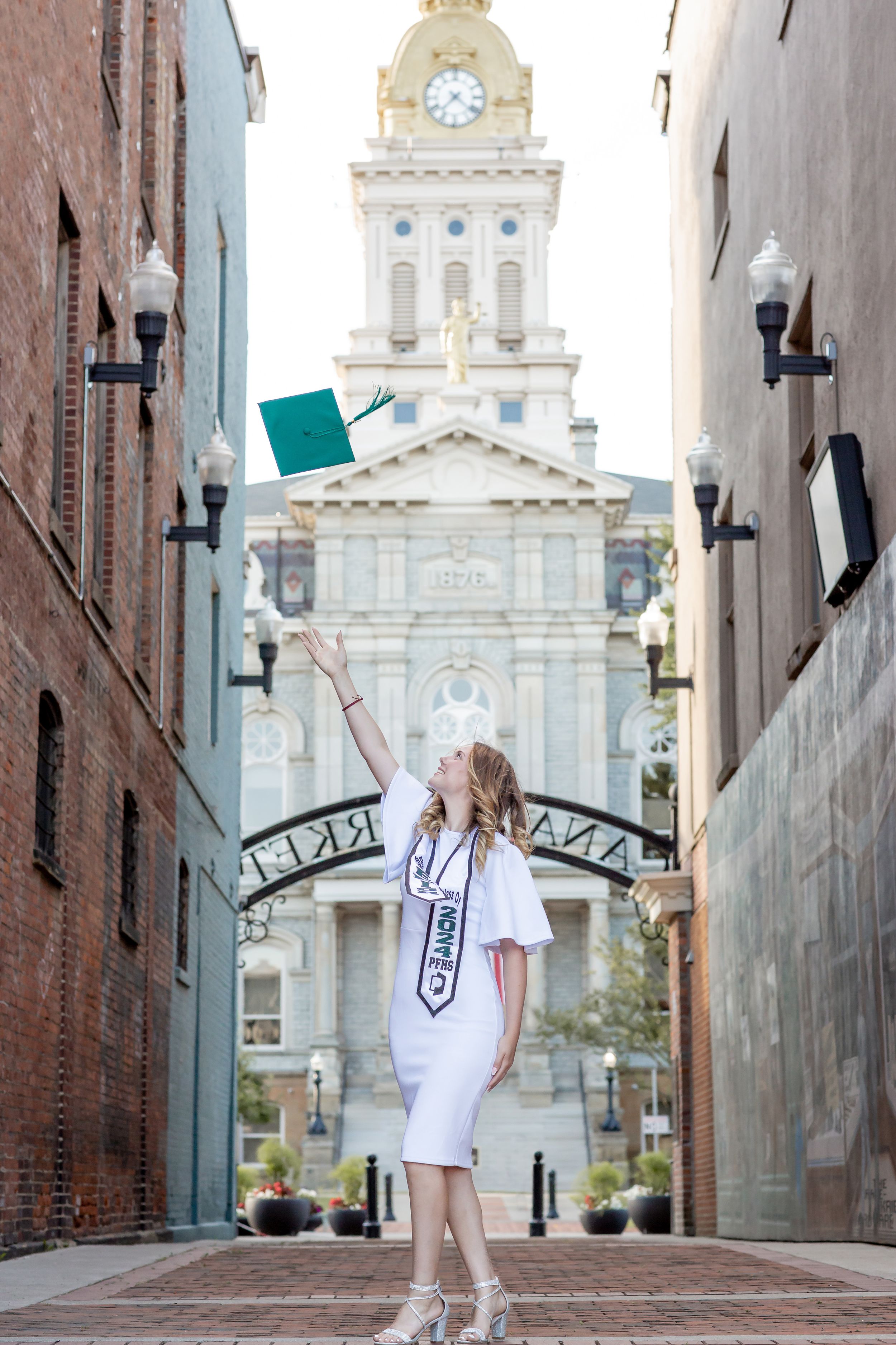 Graduate in white dress tossing green cap in front of historic courthouse building.