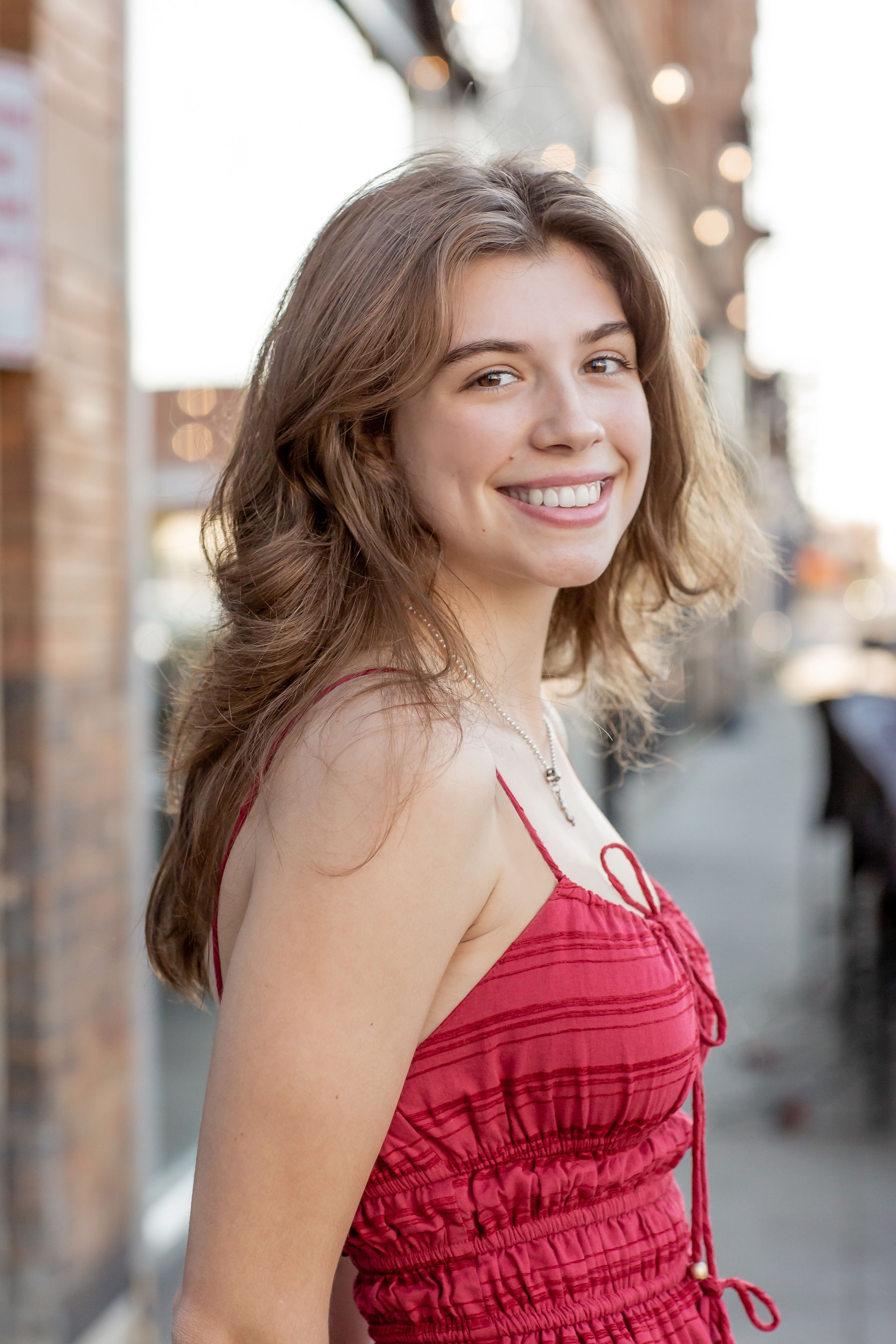 Young woman in red sundress smiling on urban sidewalk with brick building background.