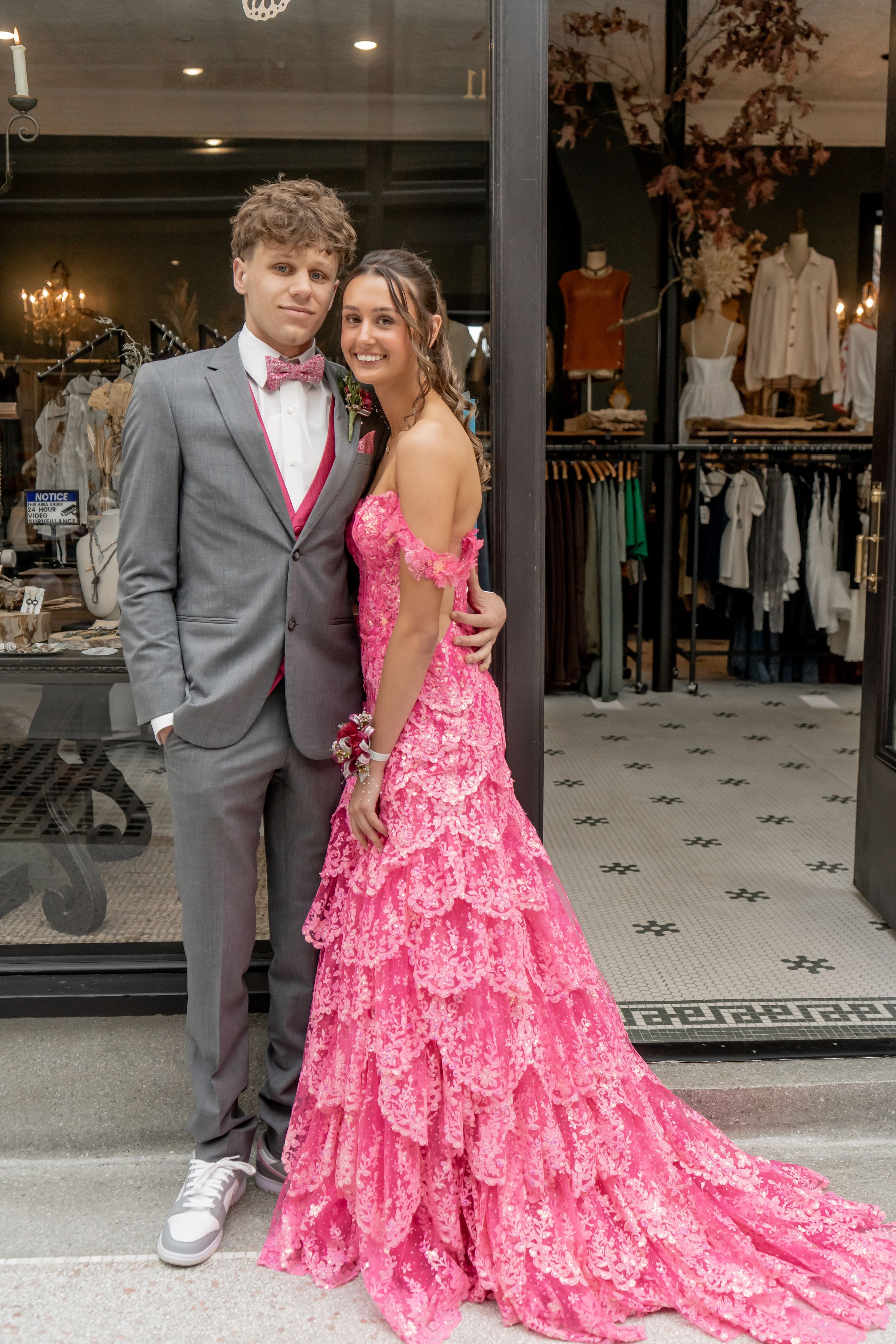 Teen couple posing in prom attire, girl in pink ruffled gown and boy in gray suit with bow tie.