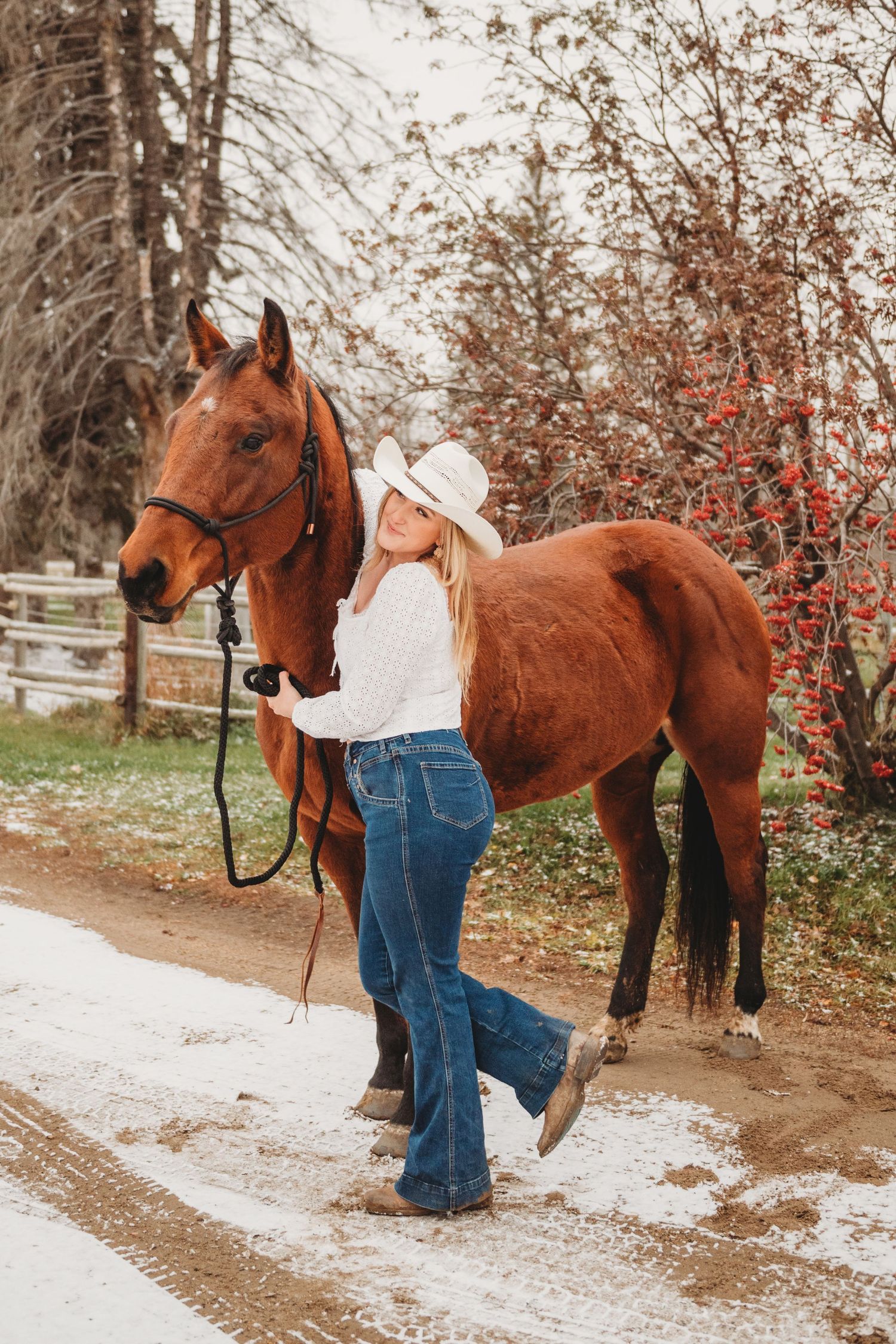 A person in western attire leads a chestnut brown horse along a snowy path bordered by autumn foliage.