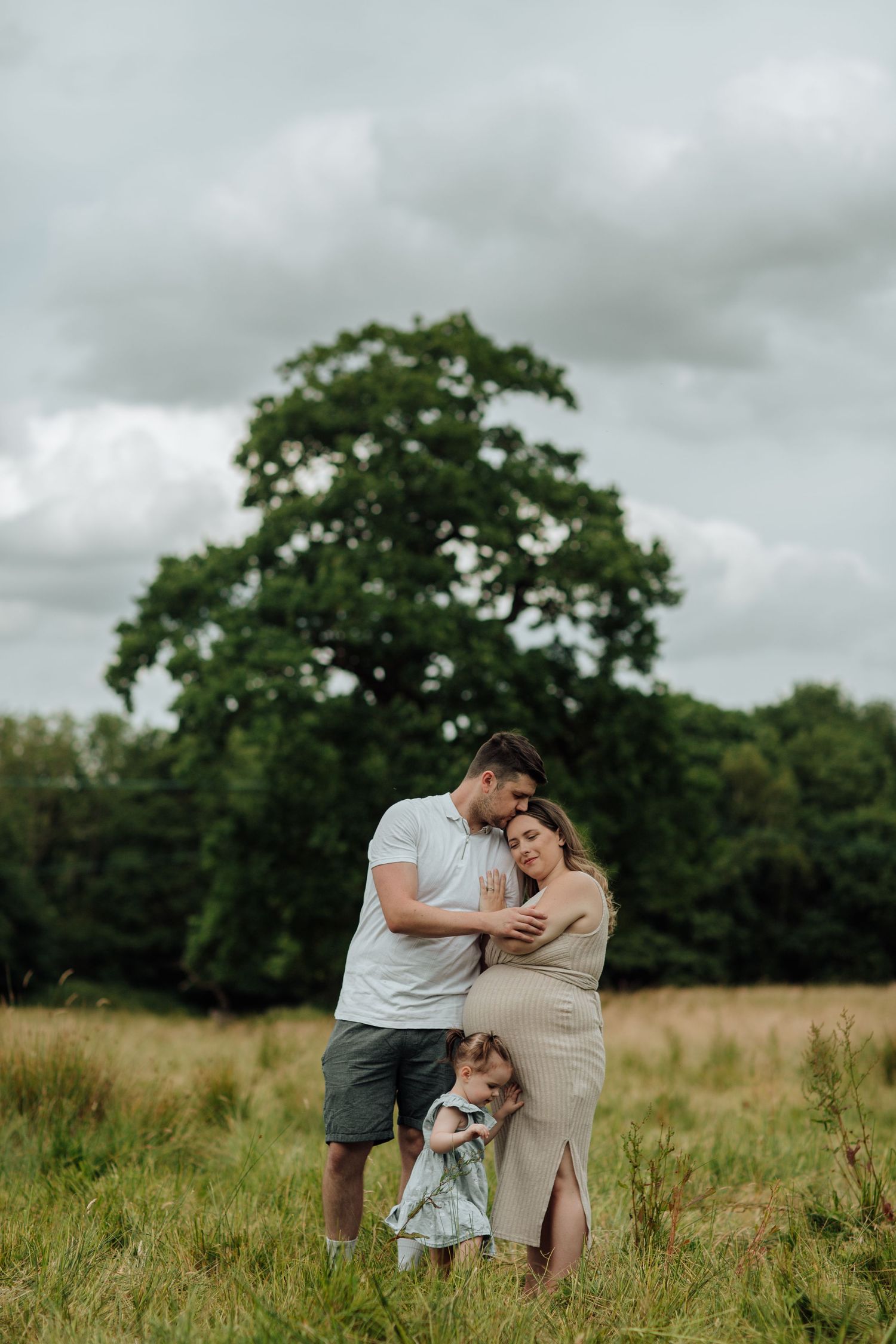 A family shares an intimate moment together while standing in a grassy meadow with a large oak tree in the background.