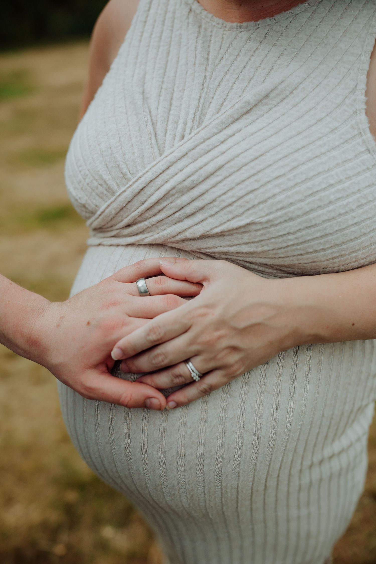 Close-up of hands with wedding rings resting on a white knit maternity dress showing a pregnant belly.