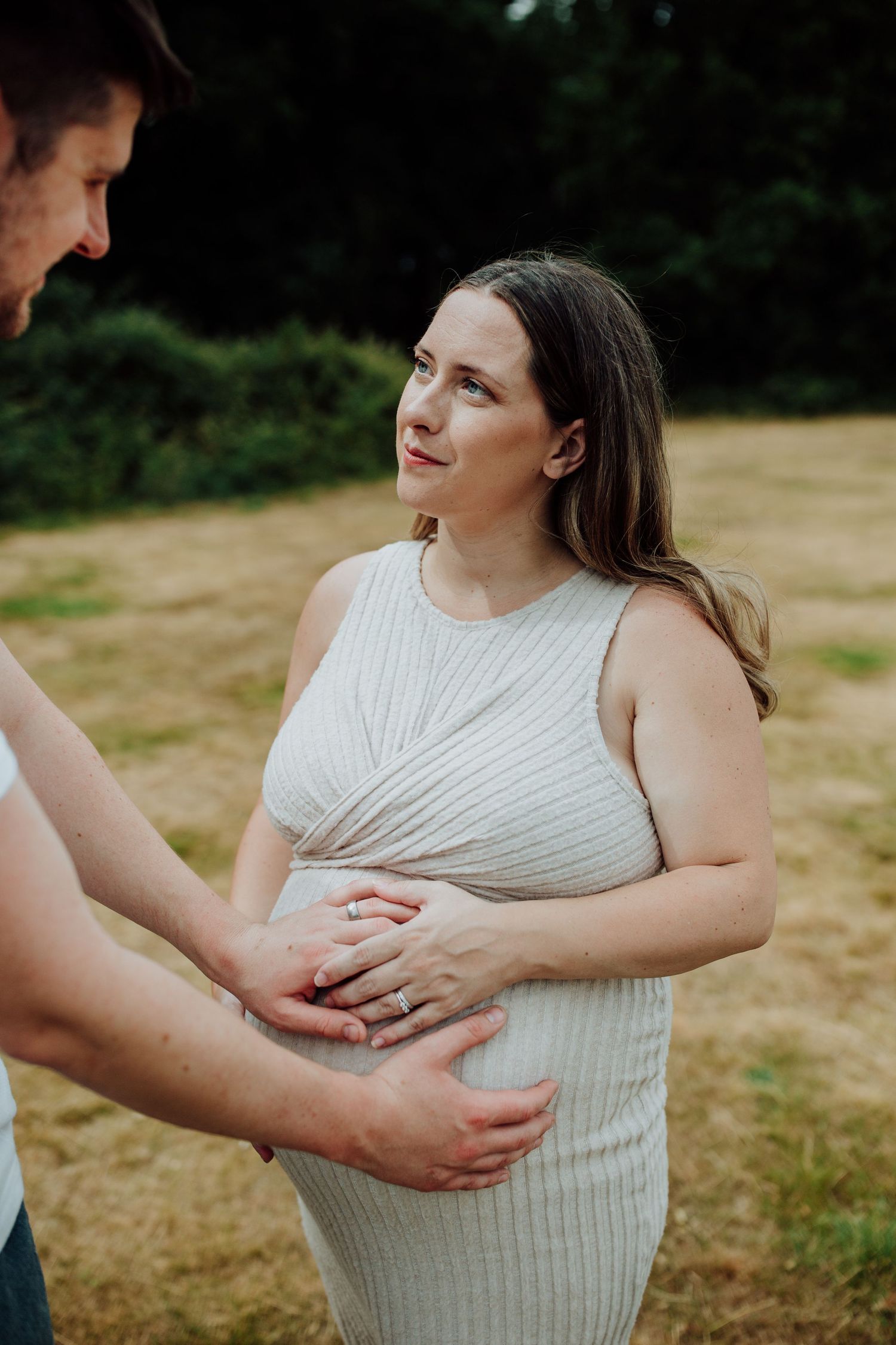 A pregnant woman in a ribbed white sleeveless dress stands outdoors while hands gently rest on her growing belly.