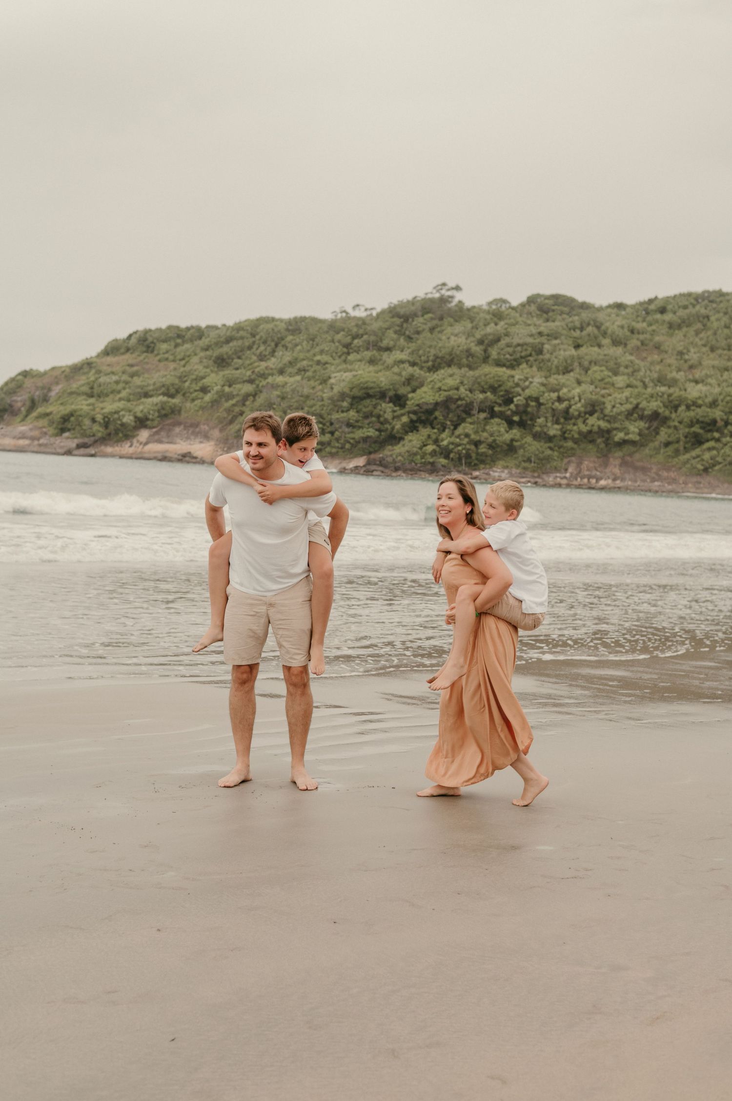 Family enjoying a beach day together, carrying children while walking on wet sand near green hills.