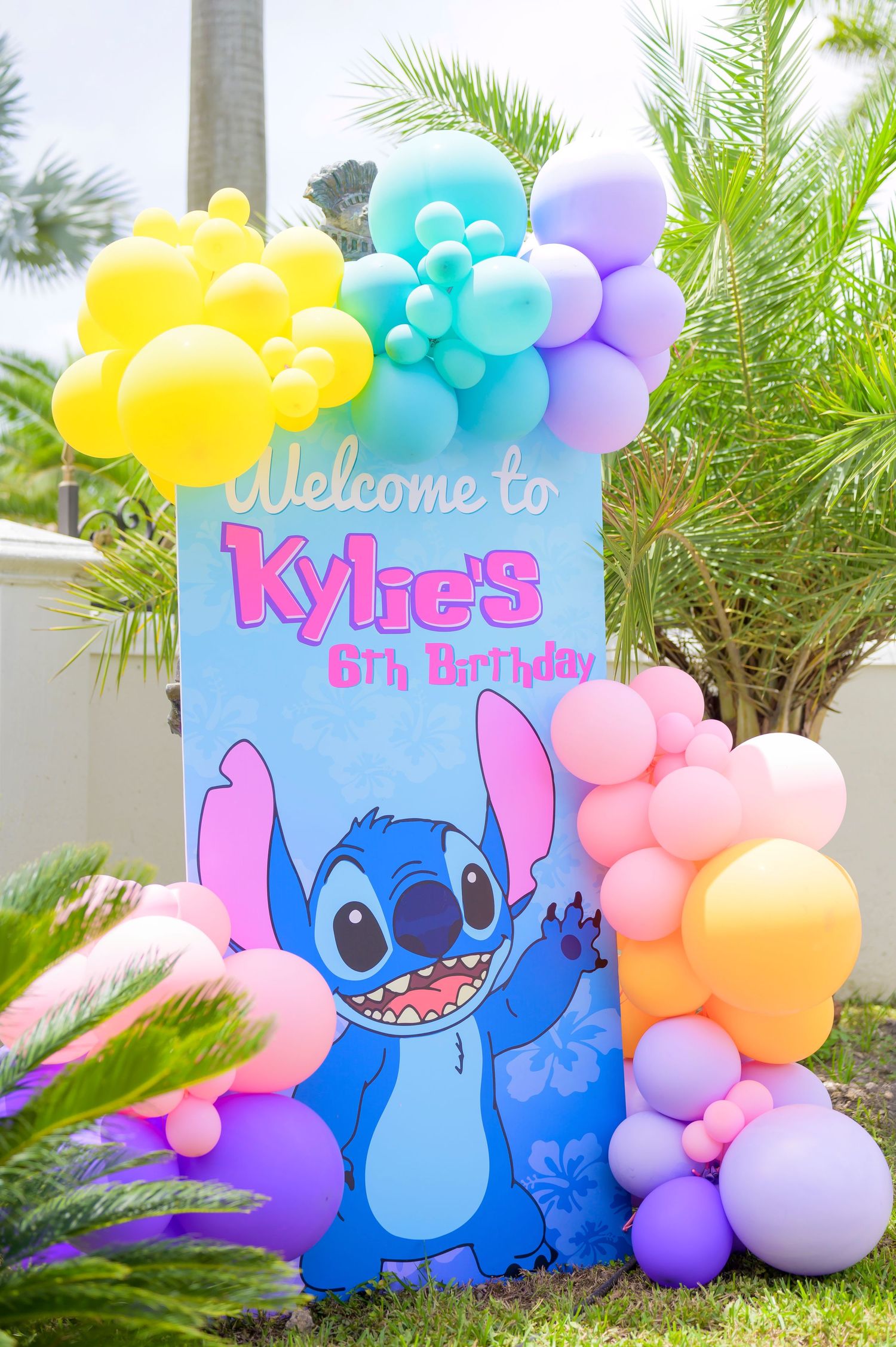 A colorful birthday party sign featuring Stitch from Lilo & Stitch decorated with pastel balloon garlands and palm trees.