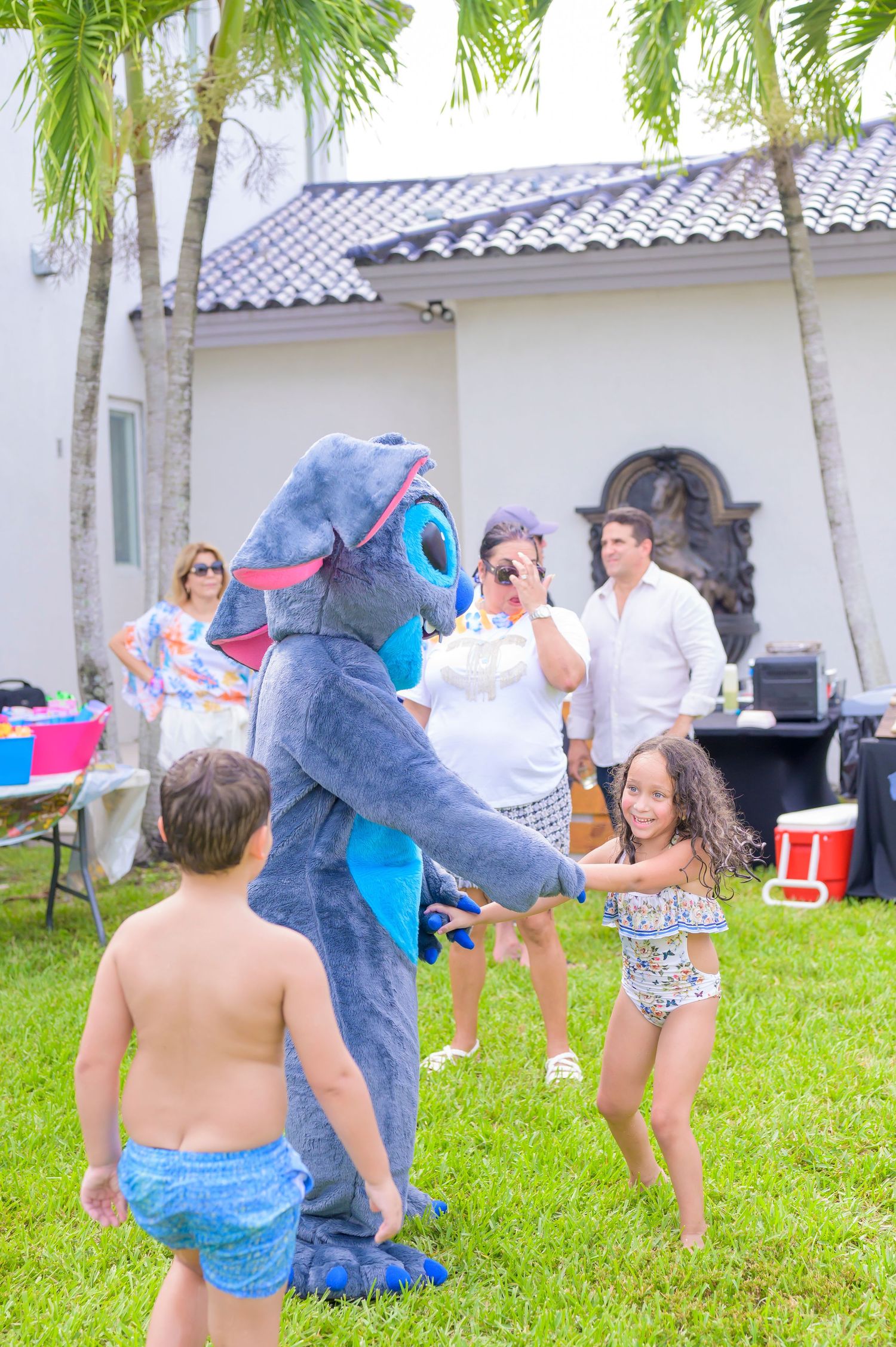 Stitch character mascot dances with kids at an outdoor party with palm trees and a white building in the background.