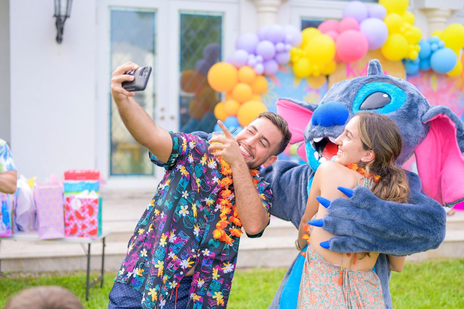 Two people pose playfully with Stitch outside a house decorated with colorful balloons for a celebration.