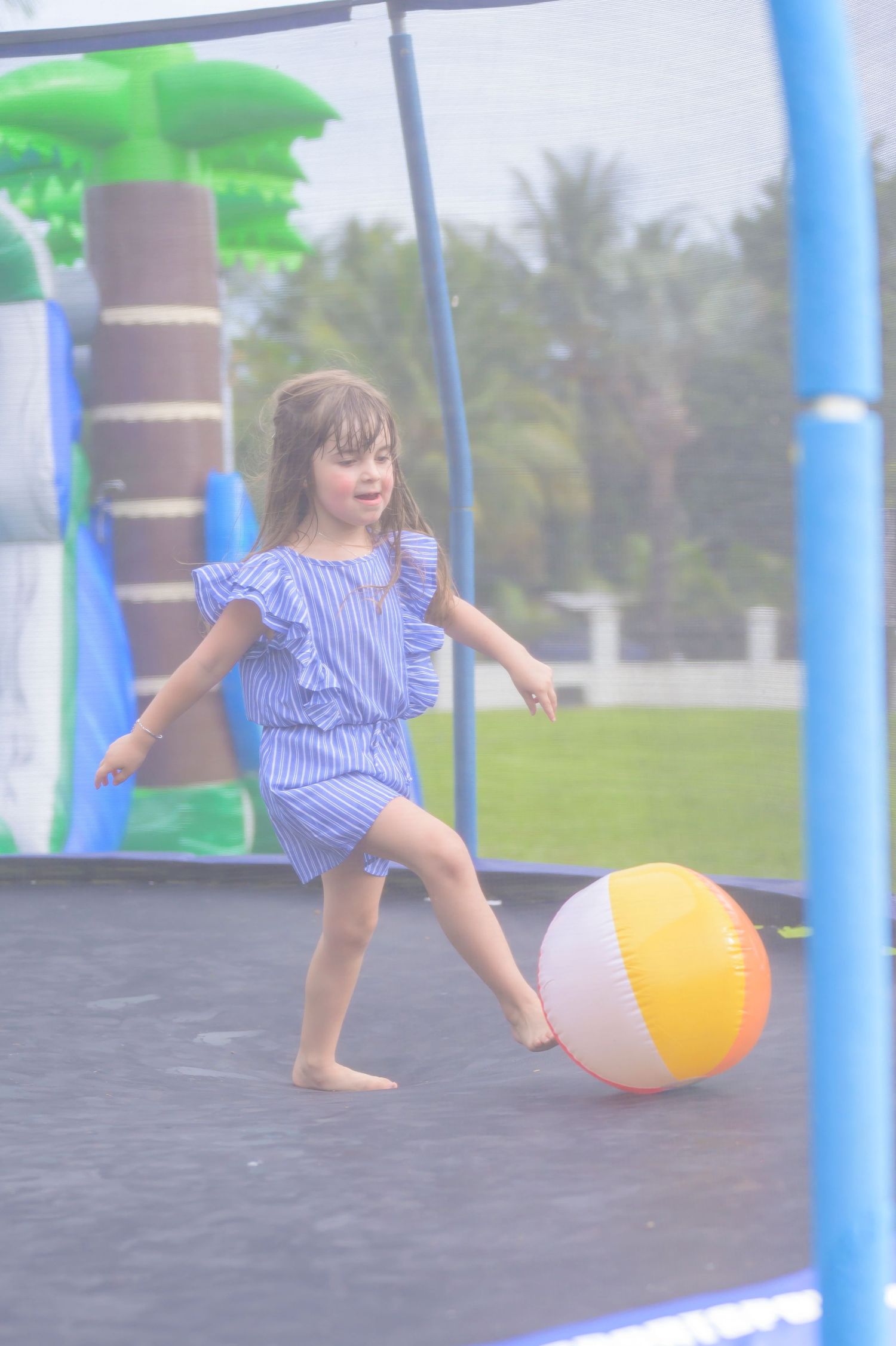 Child playing with a colorful beach ball on an outdoor playground with palm trees in the background.