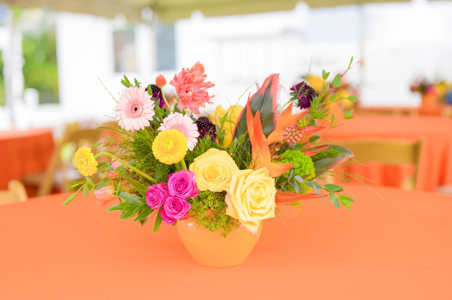 Bright floral centerpiece with pink, yellow and orange blooms arranged in a vase on an orange tablecloth.