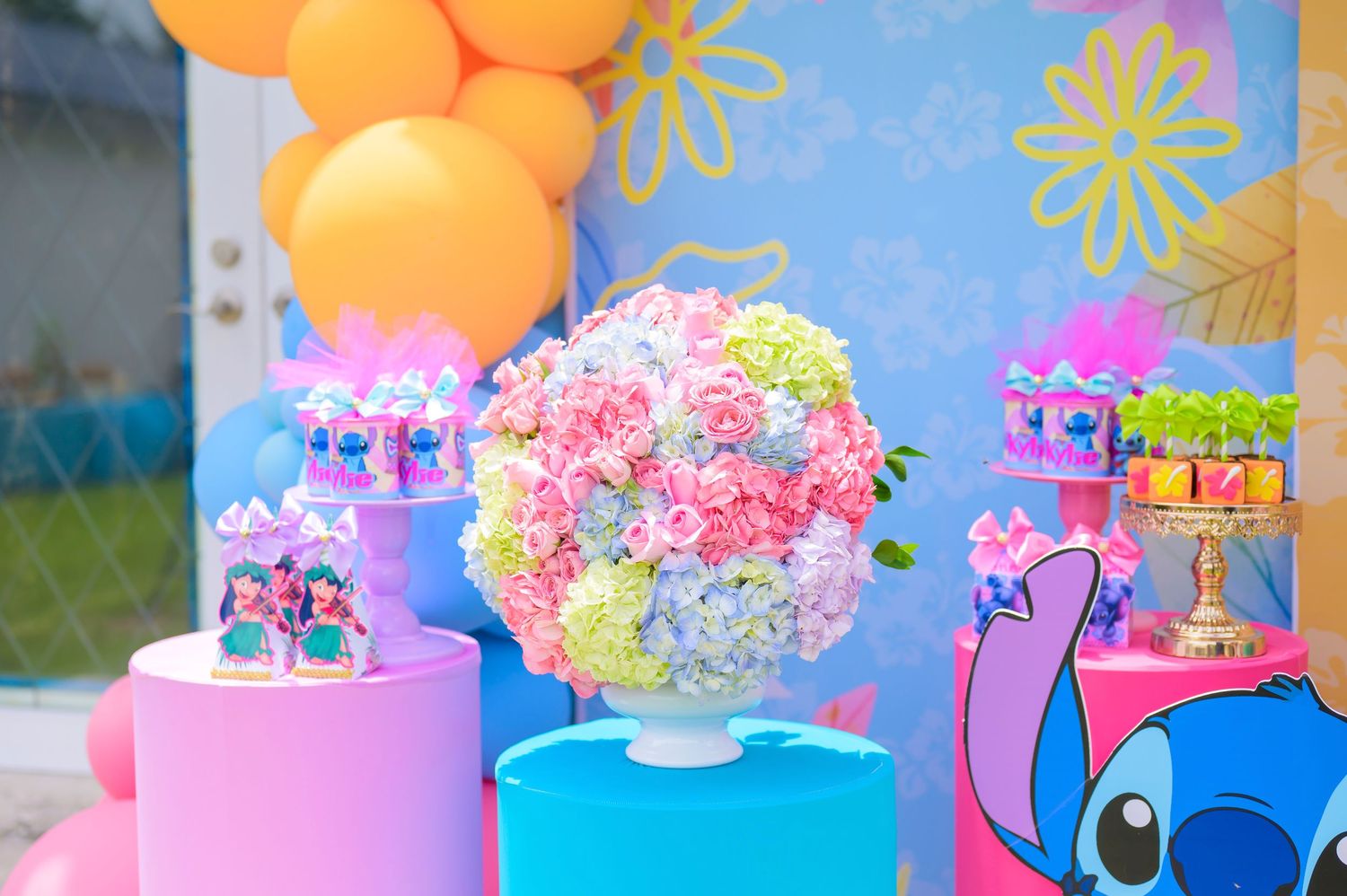 Colorful party decorations featuring pink and blue platforms with orange balloons and floral arrangements for a Stitch-themed event.