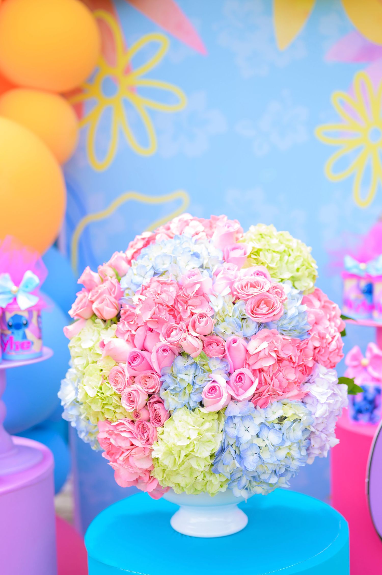 A pastel floral arrangement featuring pink roses, blue hydrangeas and yellow blooms on a turquoise pedestal against a festive backdrop.