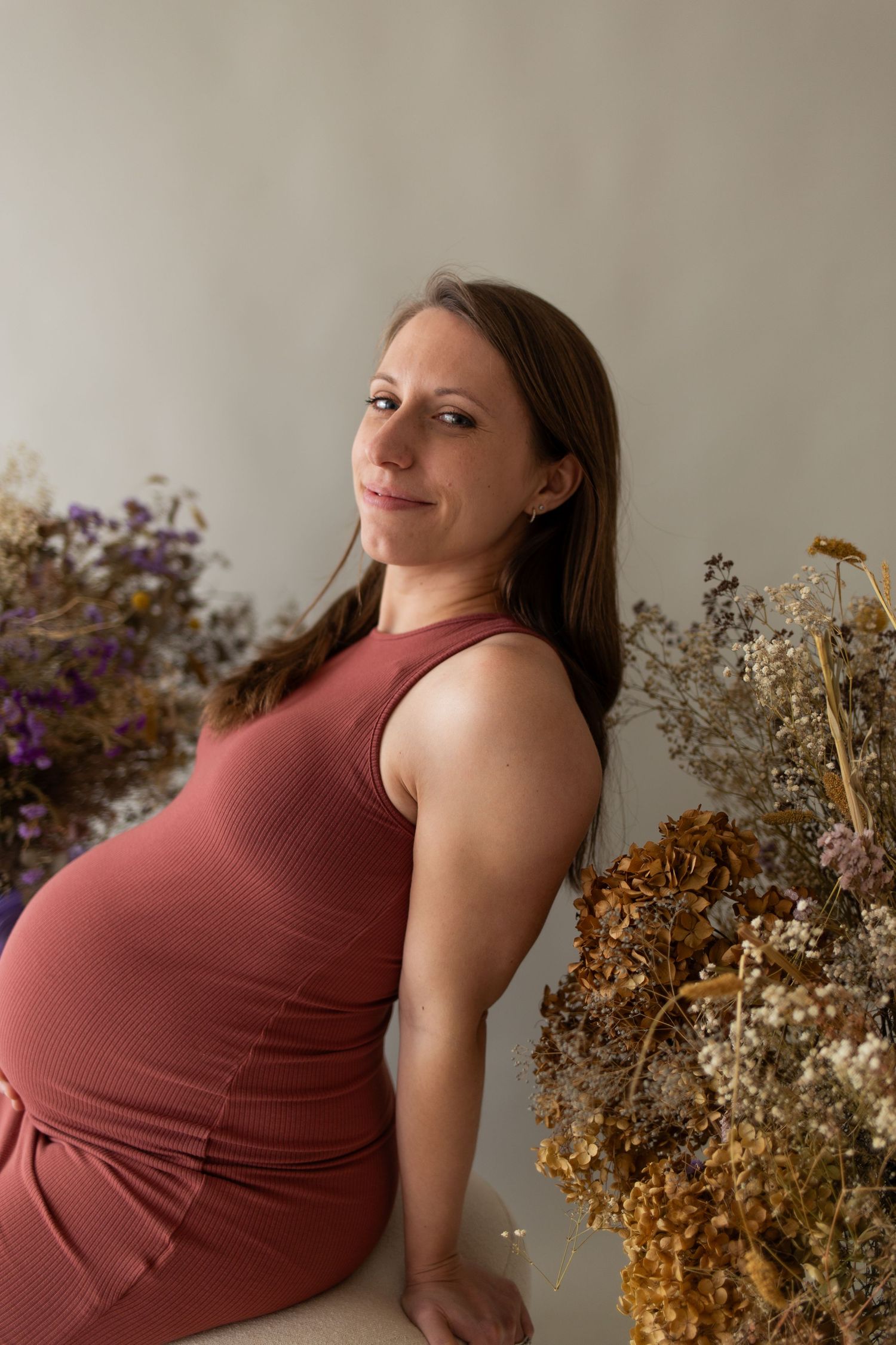 Maternity portrait of a person in a rust-colored dress near dried floral arrangements.