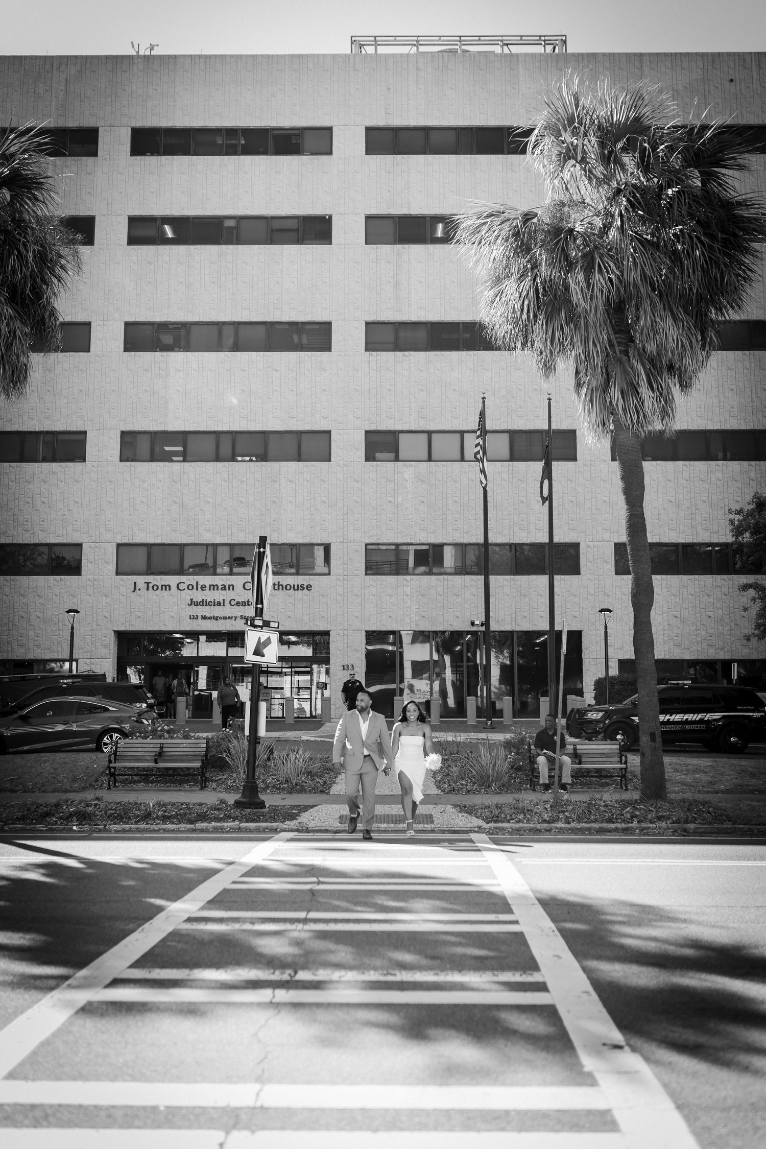 Black and white urban scene with palm tree and newlyweds walking in front of a striped building facade.