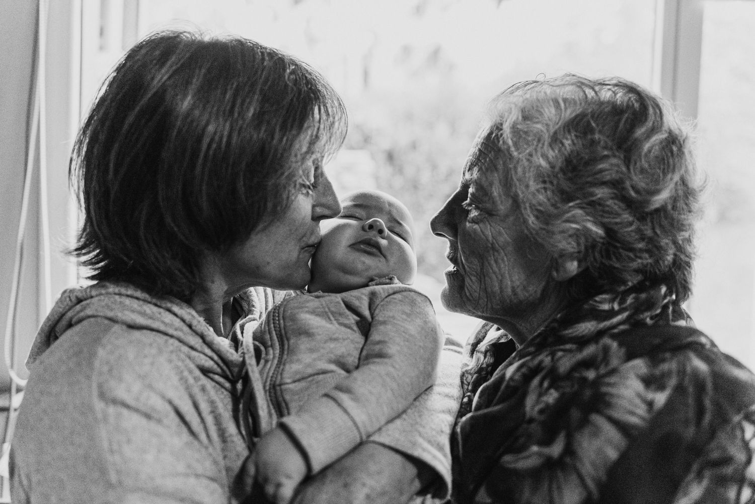 bebé recién nacido en brazos de sus abuela, ella le da un beso y la bisabuela asoma y lo mira desde el costado