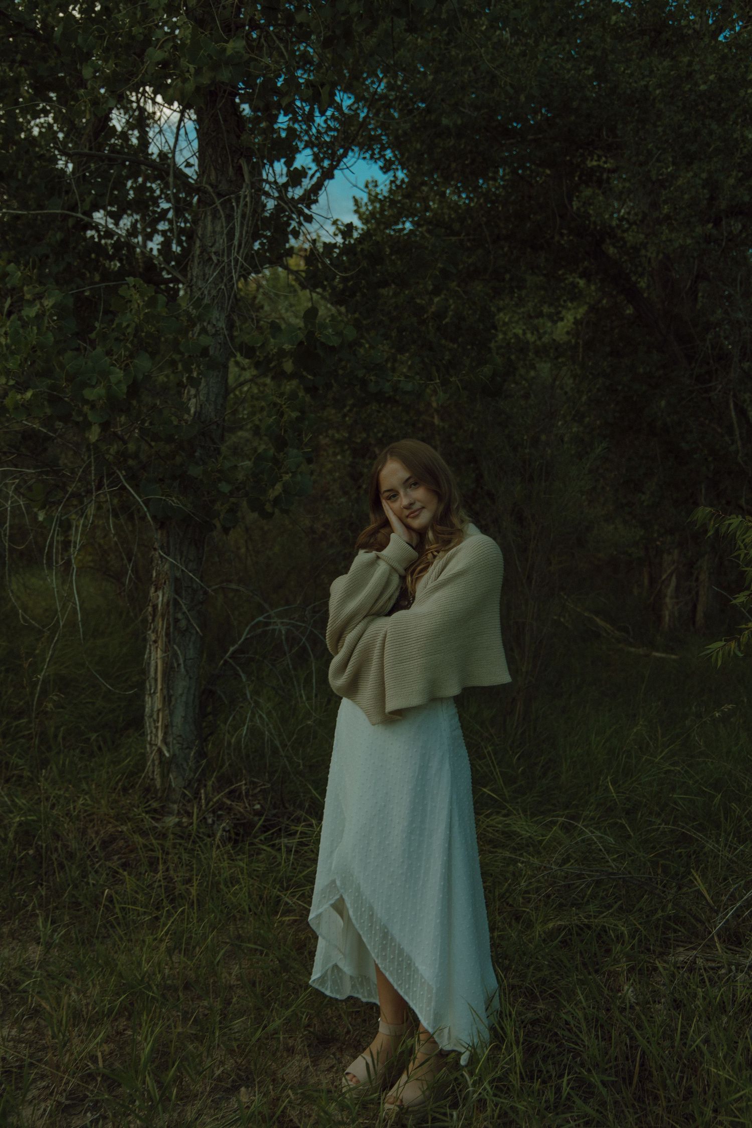 High school aged girl wearing a cardigan standing in a dark bunch of trees