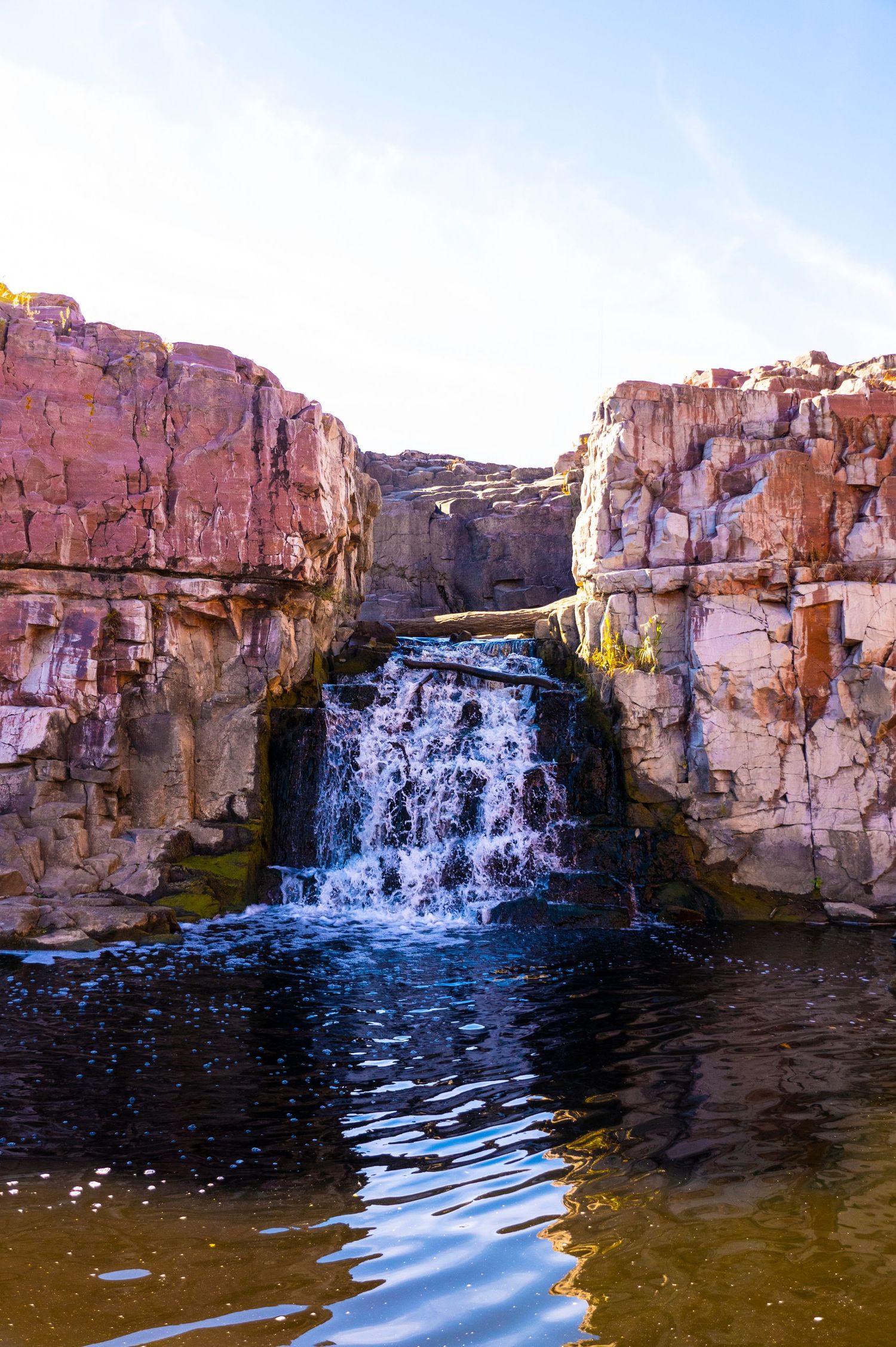 Sunlit waterfall flows through pink stone canyon walls into reflective waters below.