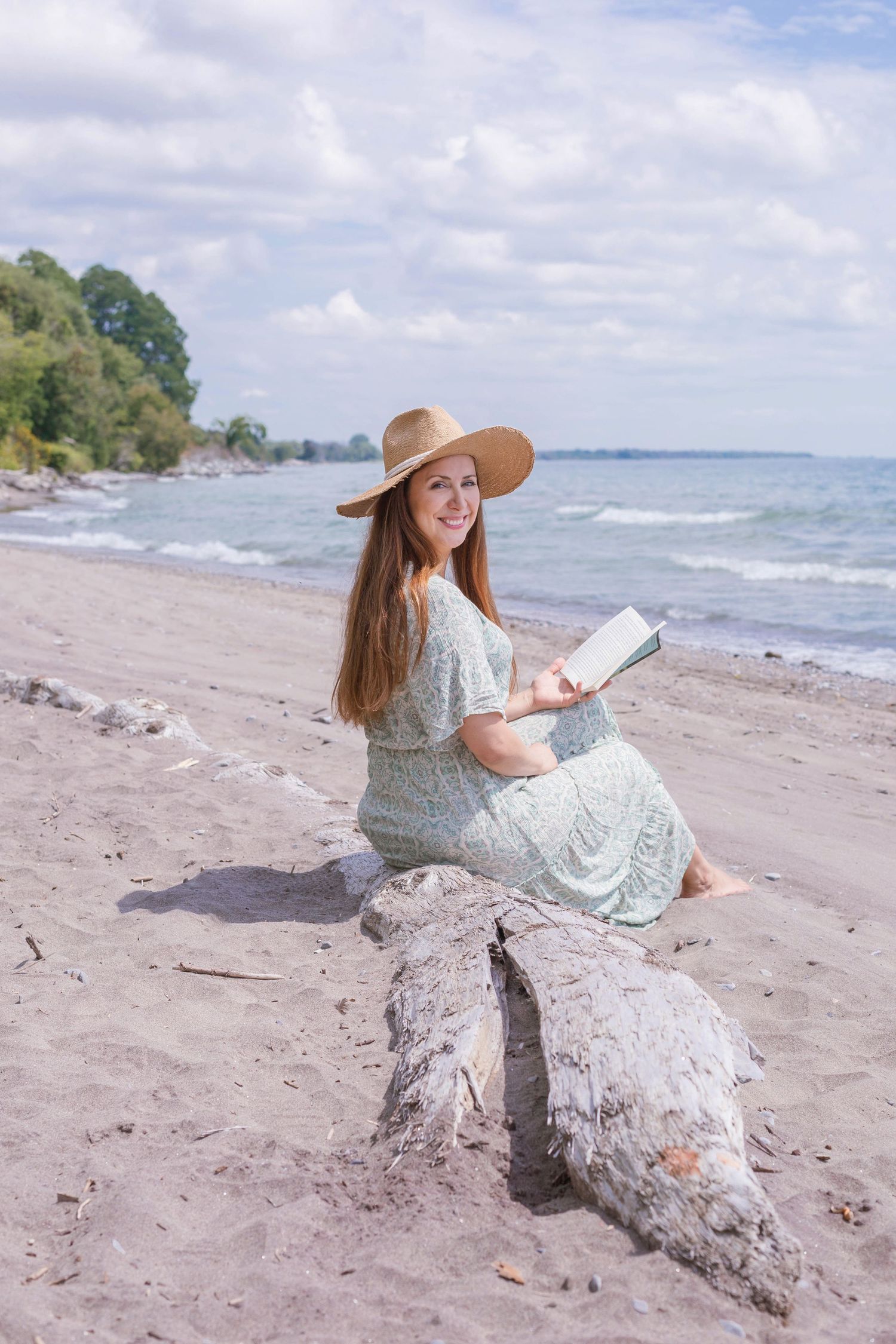 Woman in sunhat reads book while sitting on driftwood at scenic beach.
