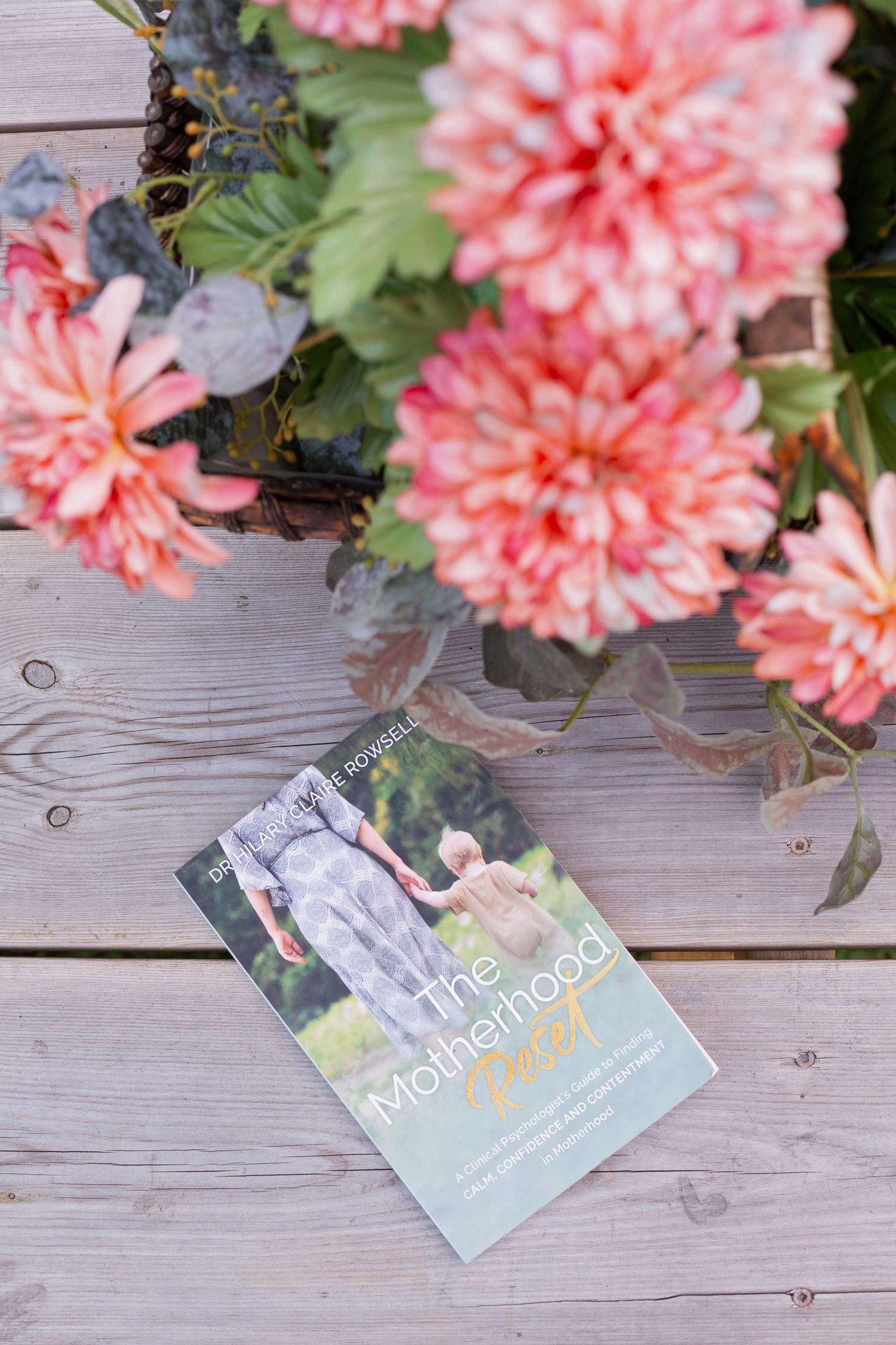 Book displayed on wooden deck with coral-colored dahlias in foreground.
