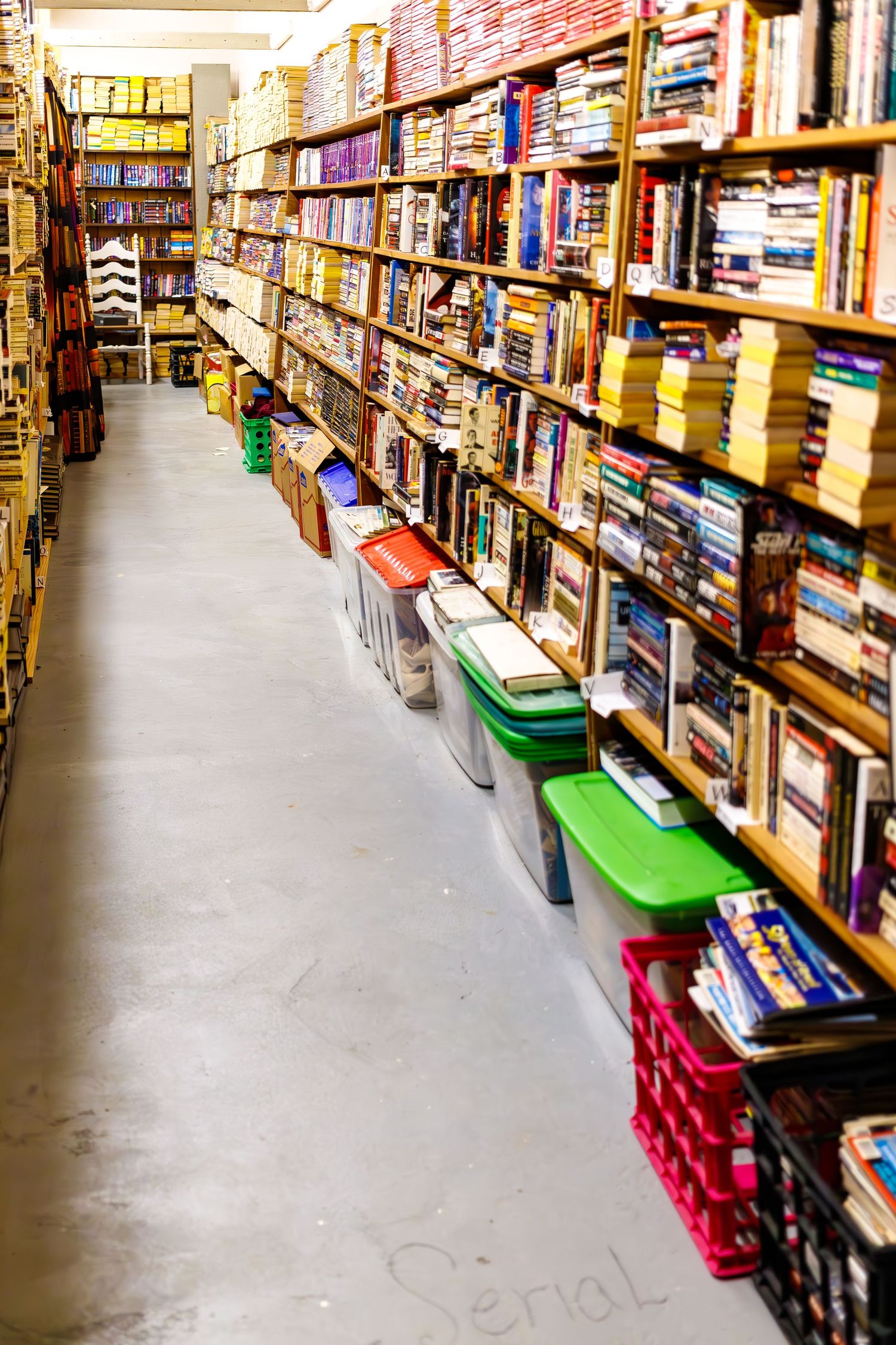 Long aisle of packed bookshelves in a bookstore with storage bins along the floor.