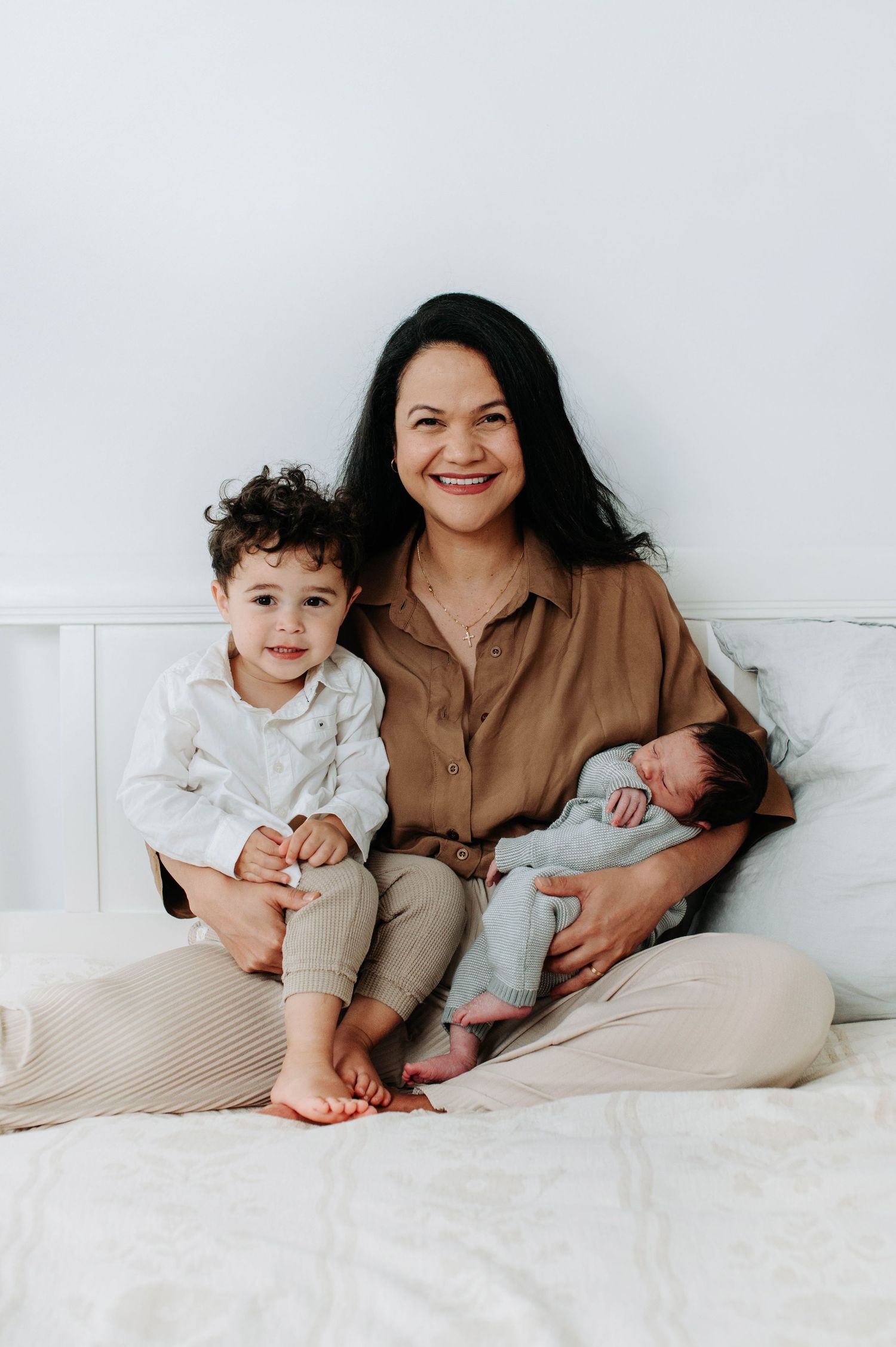 A smiling mother sits on a white bed holding a newborn baby while her young toddler sits beside her.