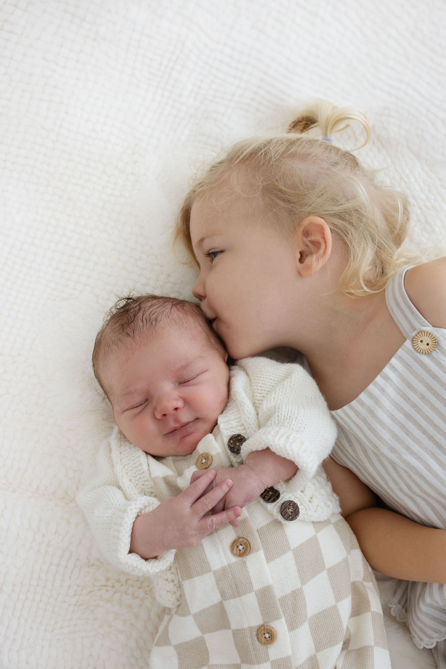 A toddler girl with blonde hair gently kisses a sleeping newborn baby wrapped in a checkered knit blanket.