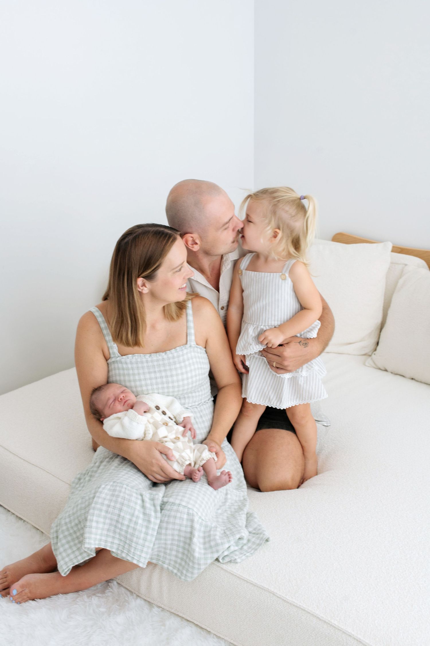 A family sits on a white couch, mother holding newborn, father kissing toddler daughter in a bright minimalist room.