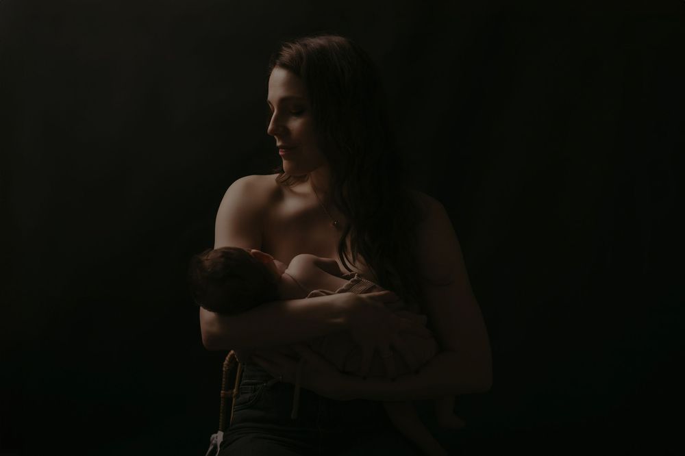 A mother tenderly holds her newborn baby while breastfeeding in a dramatic dark studio portrait.