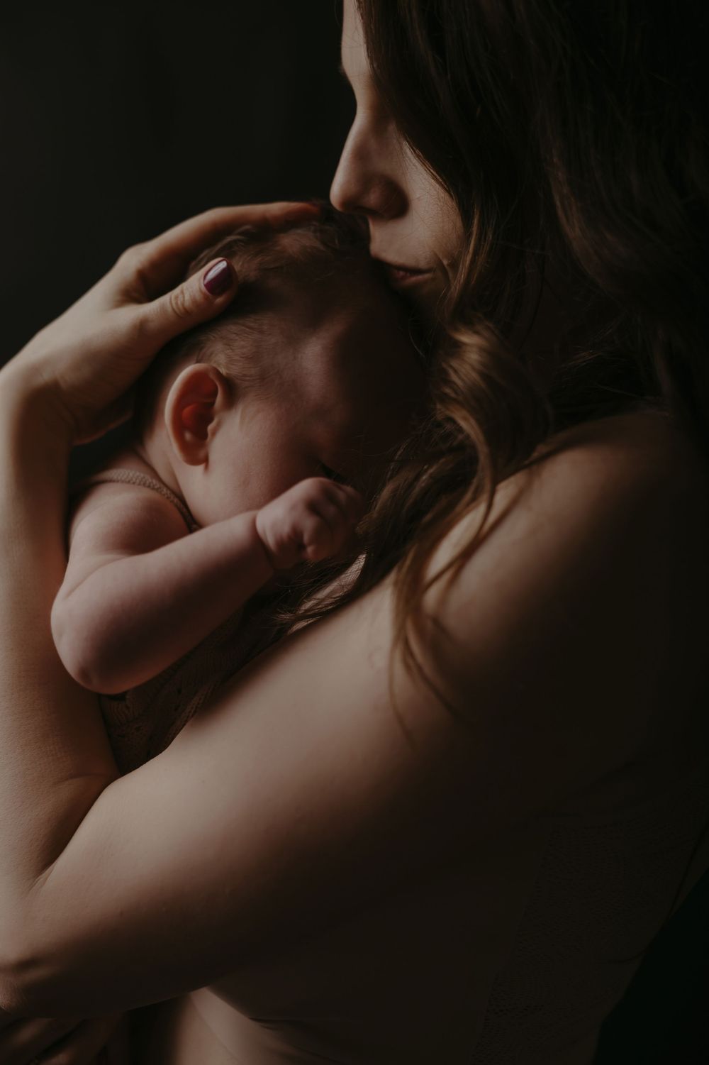 A mother tenderly kisses her newborn baby while cradling them against her chest in soft, dark lighting.
