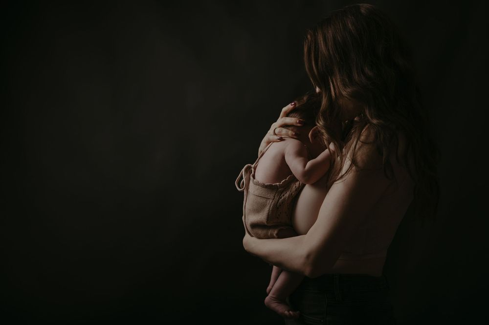 A mother tenderly holds her newborn baby against a dark, moody studio background in an intimate portrait.