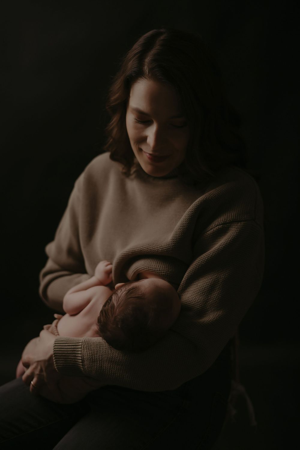 A smiling mother in a beige sweater gently holds her sleeping newborn baby against a dark background.