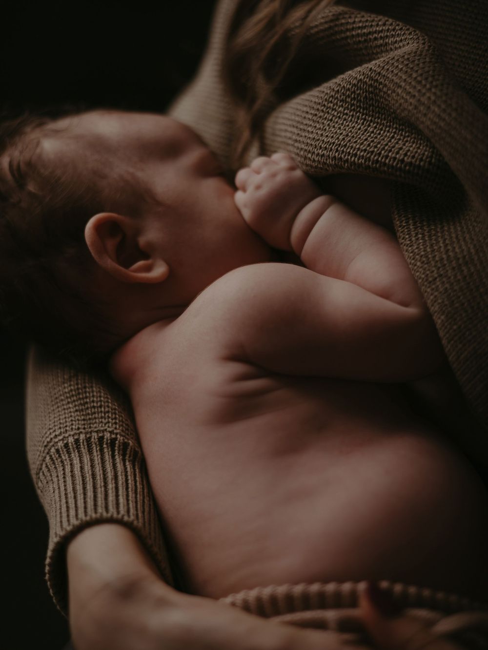 Close-up of a newborn baby nursing, cradled in a mothers arms wearing a cozy knit sweater.