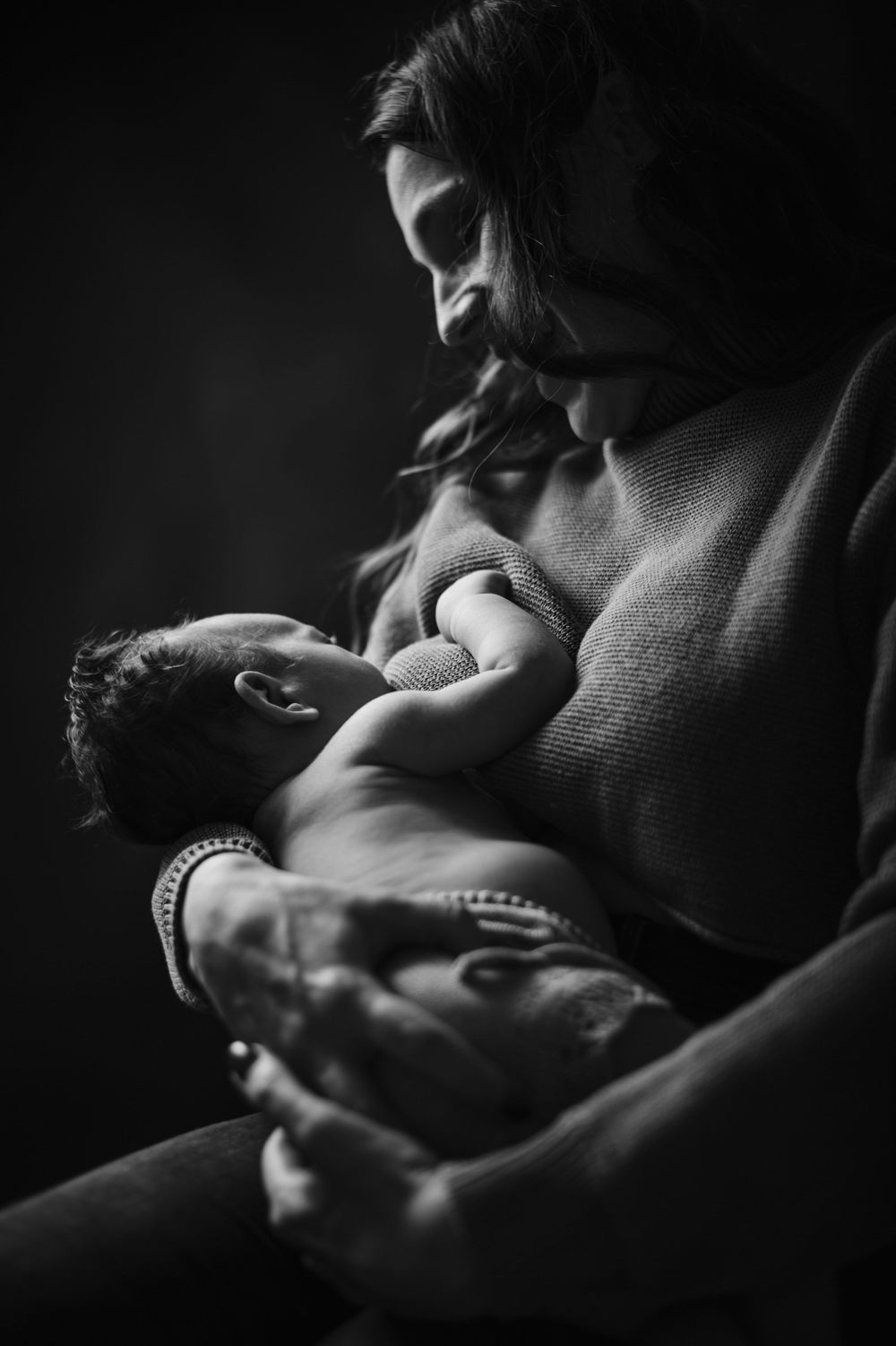 Black and white photo of a mother tenderly cradling her newborn baby close to her chest in soft light.