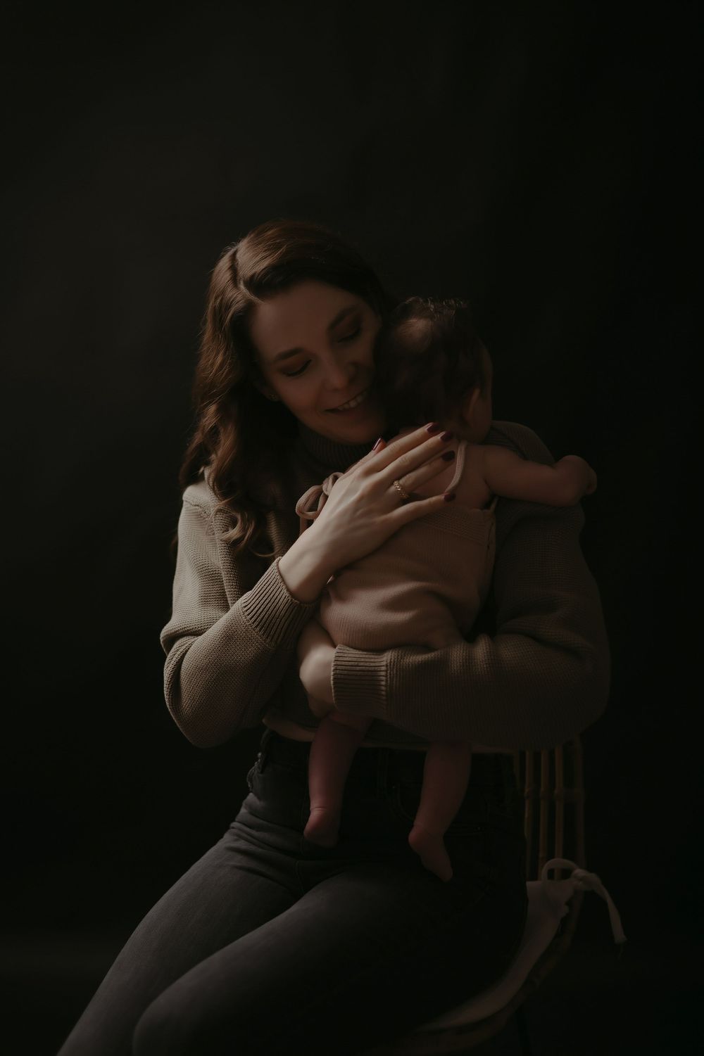 A mother tenderly holds her newborn baby while seated on a chair against a dark studio background.