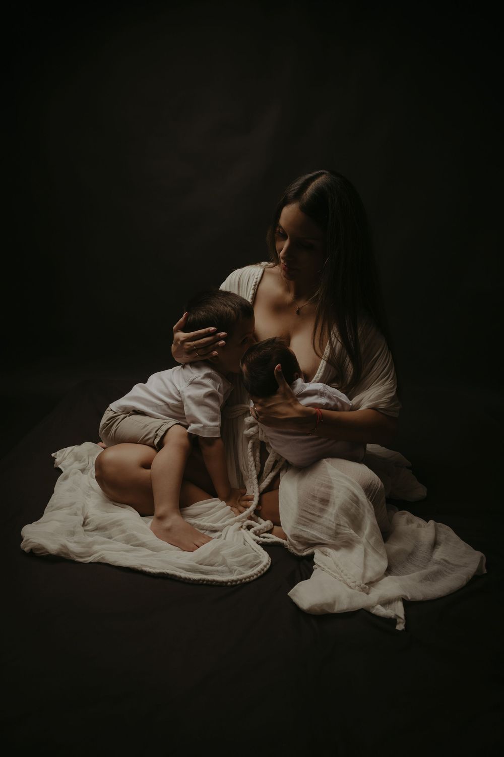 A mother in white fabric sits on the floor cradling her newborn baby in a dark, moody studio portrait.