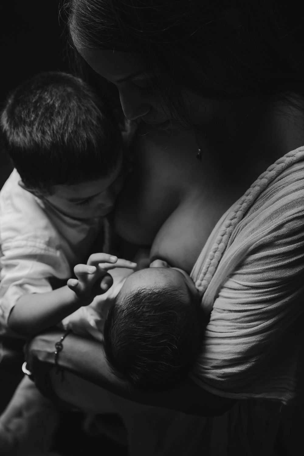 Black and white photo of a mother breastfeeding a newborn while an older toddler child looks on tenderly.